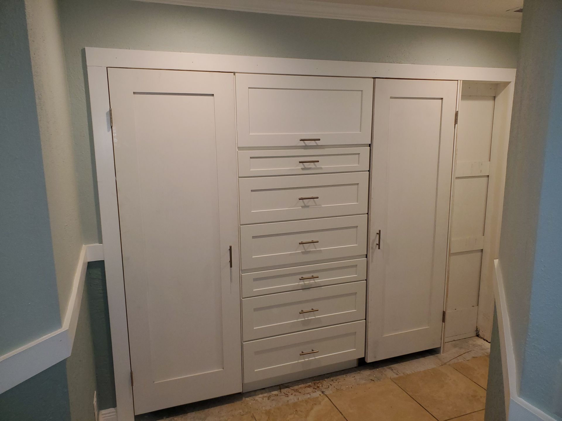 Built-in white storage unit with two tall doors flanking six central drawers against a pale green wall and tiled floor.