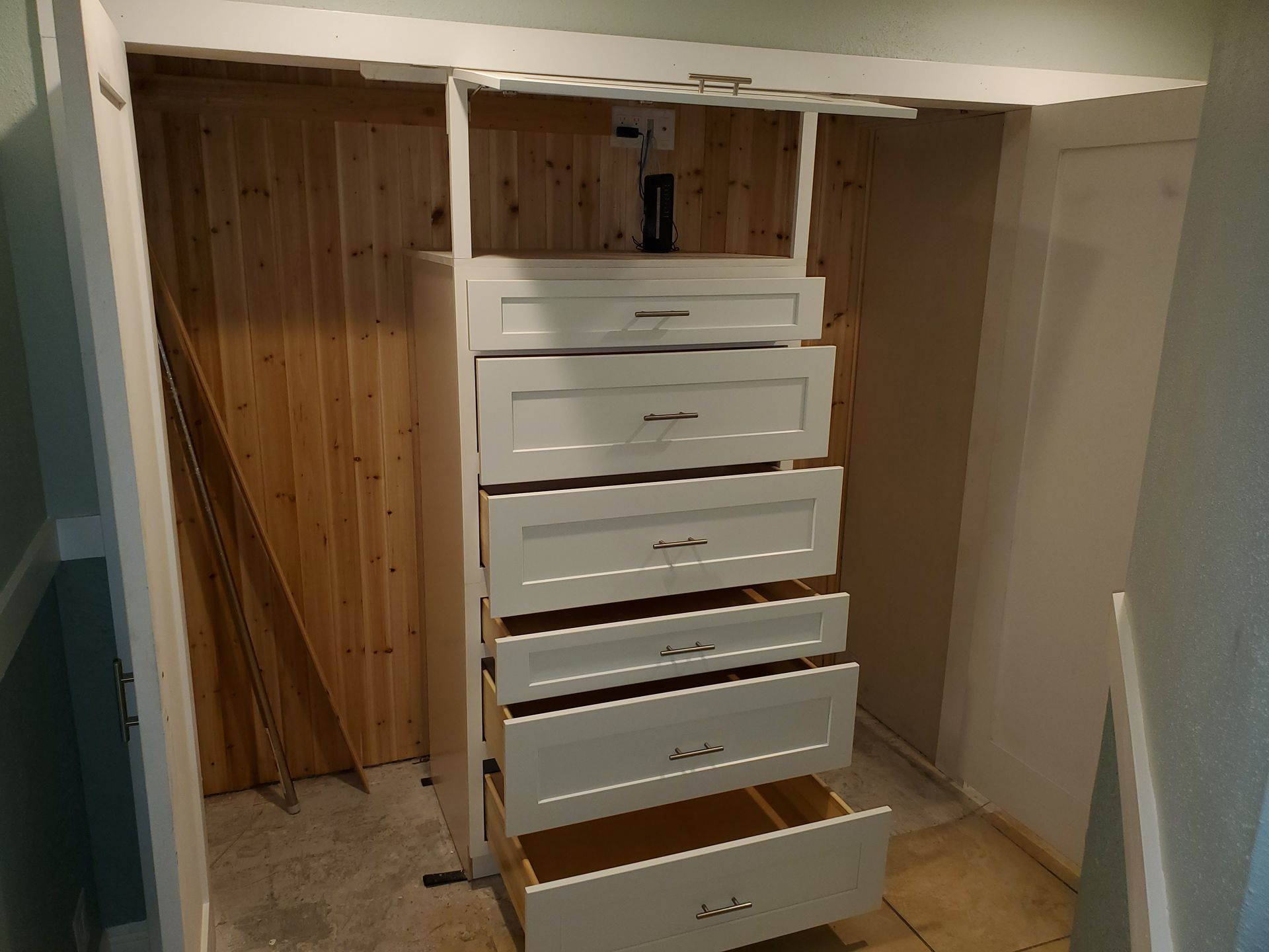 White dresser with multiple open drawers sitting inside a closet with wood-paneled walls.