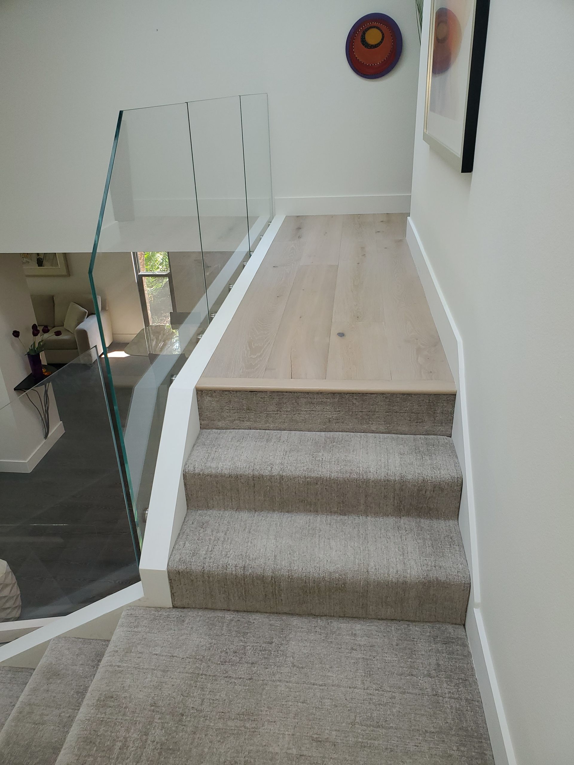 A carpeted staircase leading to a landing with light wood flooring and a modern glass railing in a bright hallway.