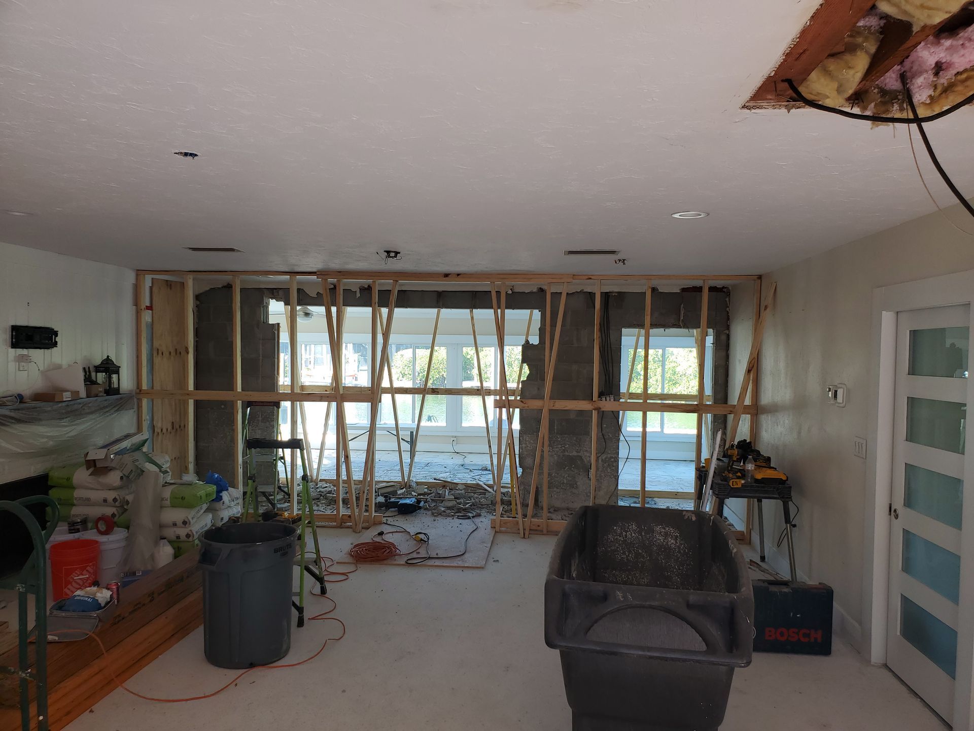 A residential interior under construction, showing a wooden framed wall opening to an outdoor view, tools, and debris.