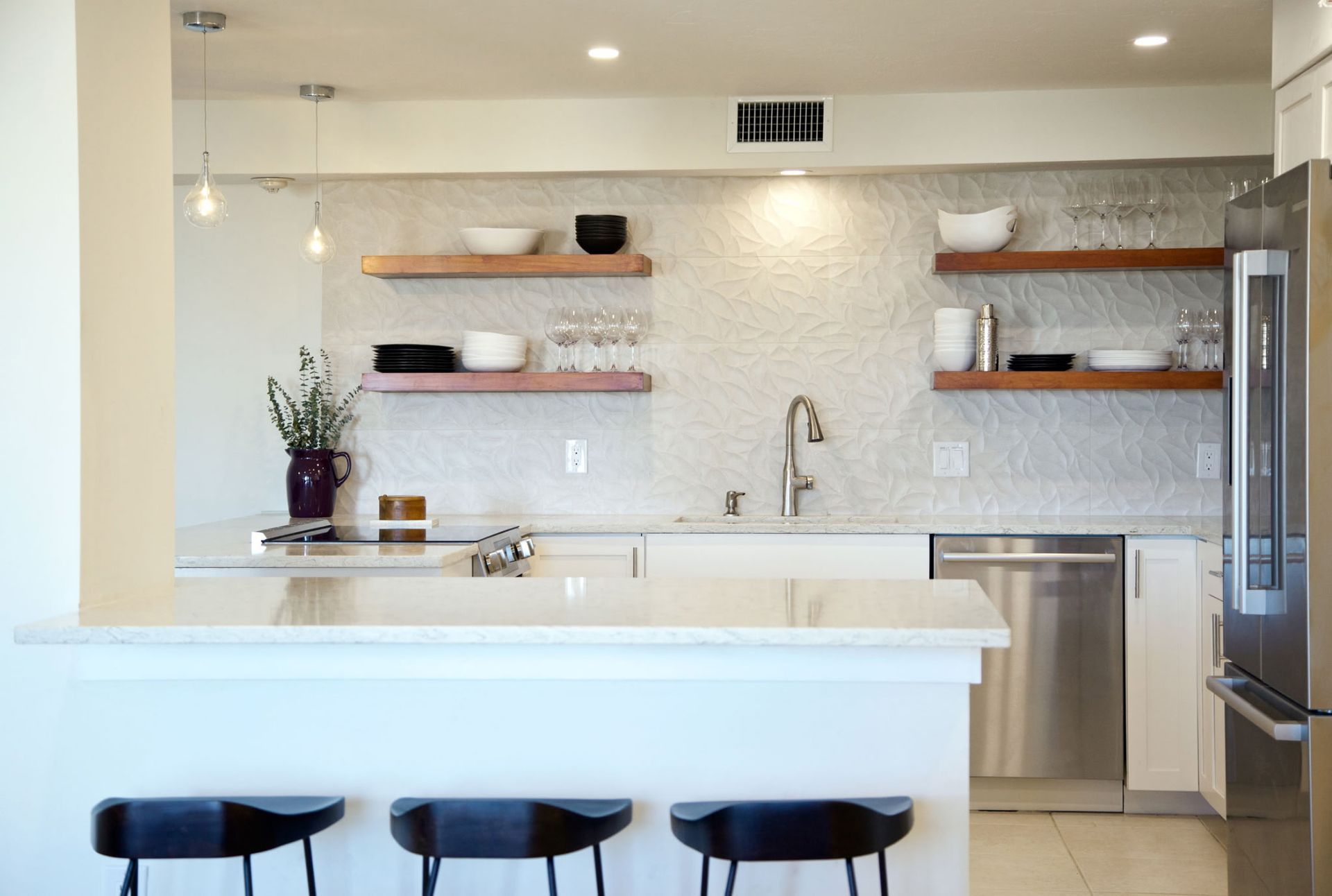 A modern kitchen with white cabinets, light countertops, open wooden shelves, and three black stools at a breakfast bar.