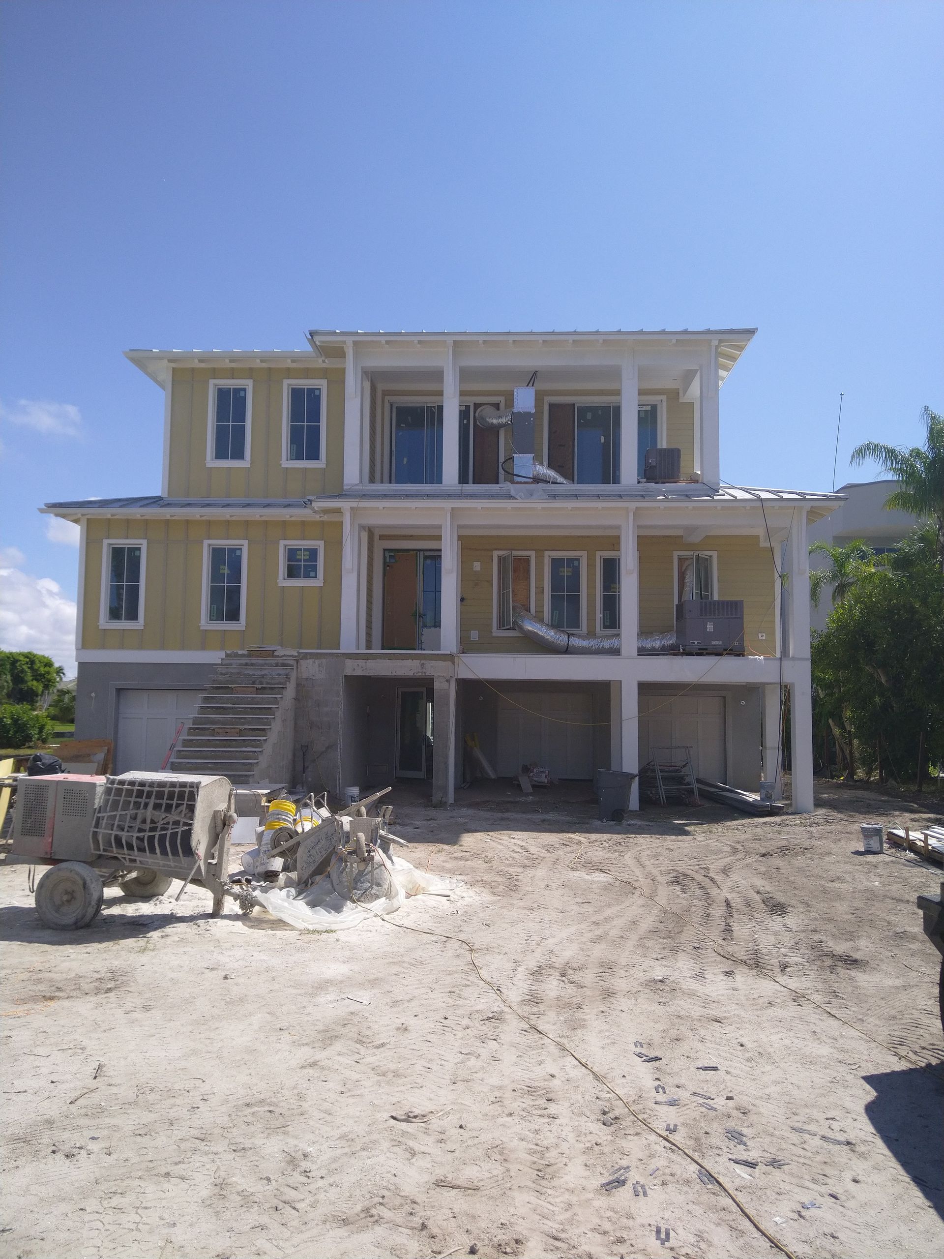 A two-story house under construction with yellow siding, a raised foundation, a porch, and sandy ground in the foreground.
