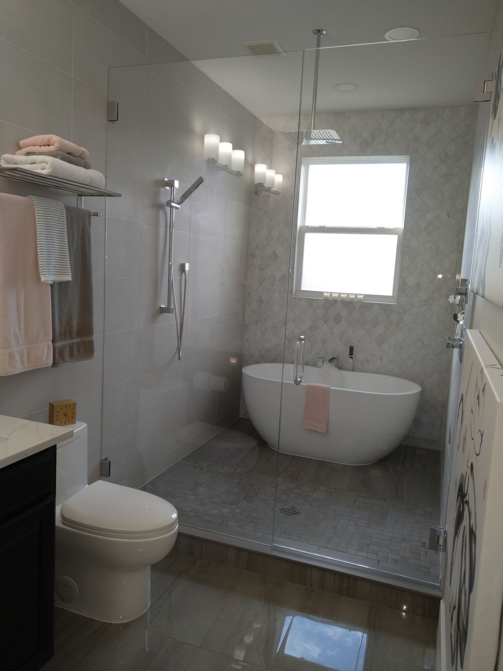 A modern bathroom with a white freestanding tub in a glass-enclosed shower area, a white toilet, and gray tiled walls.