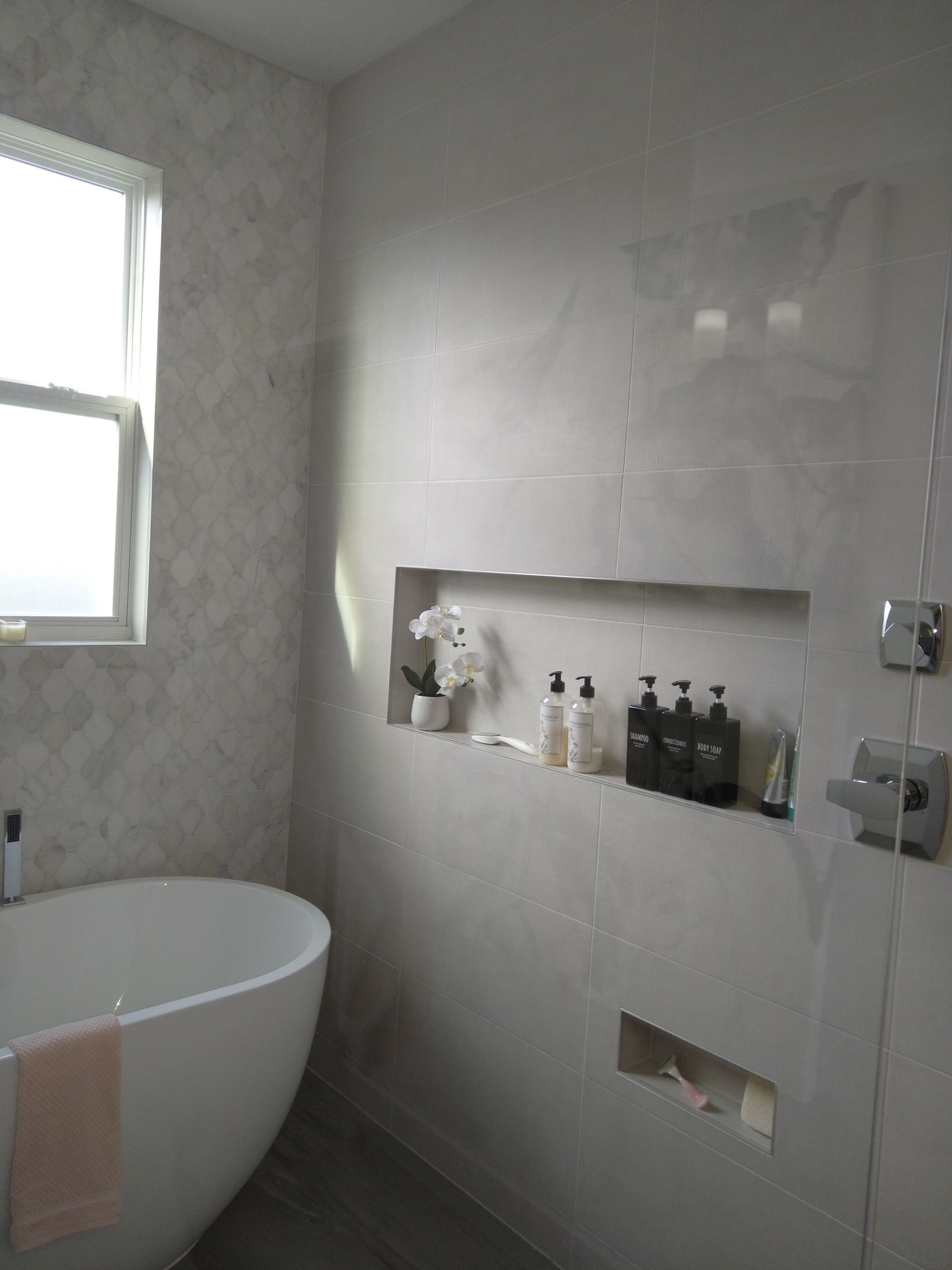 A white bathtub sits beside a tiled wall featuring a built-in recessed shelf filled with toiletries and a small plant.