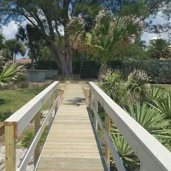 Wooden boardwalk with white railings leads toward greenery, a tree, and a building under a blue sky.