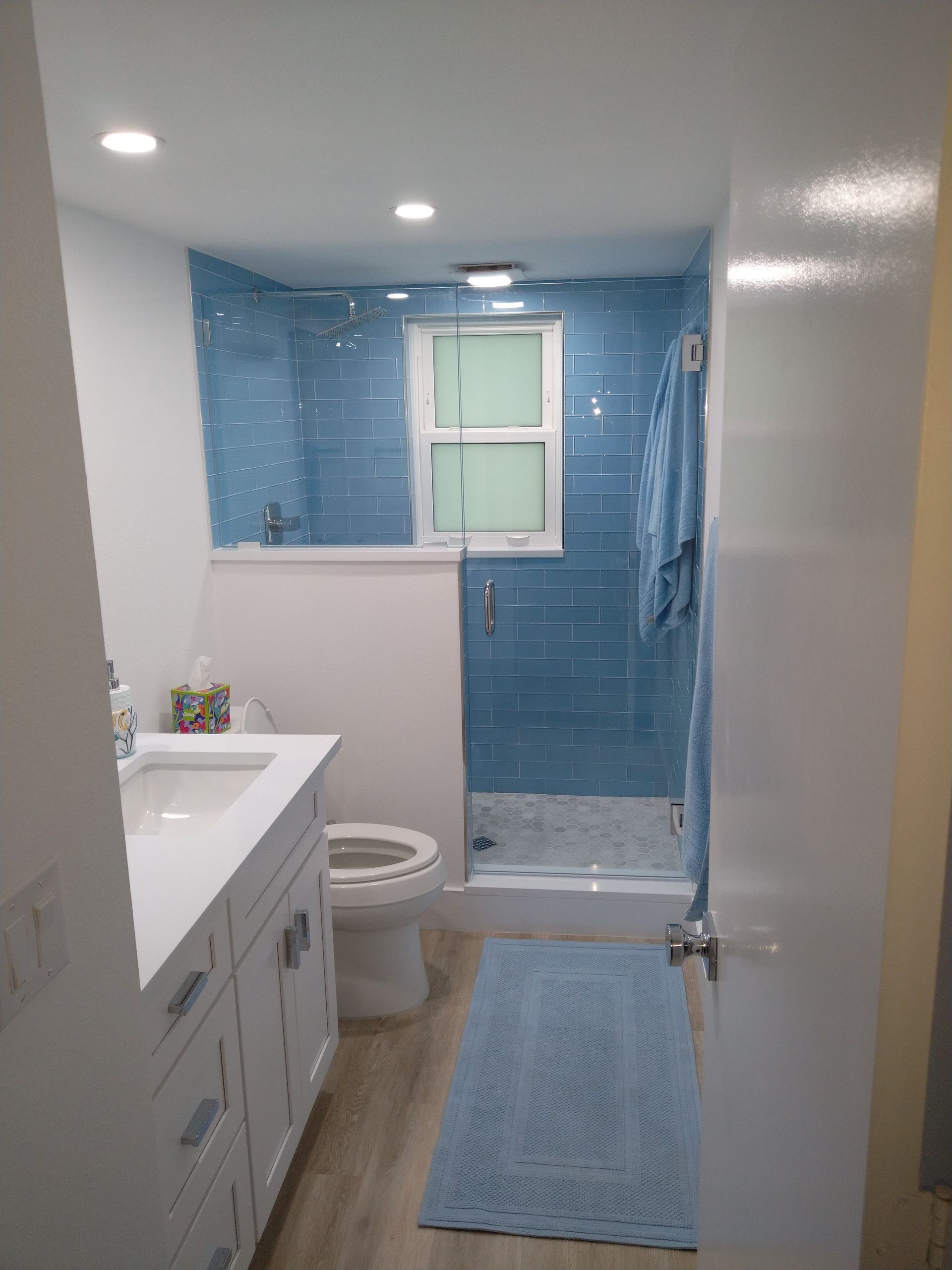 A bathroom with a white vanity, a toilet, and a walk-in shower with blue tiled walls and a light wood-look floor.
