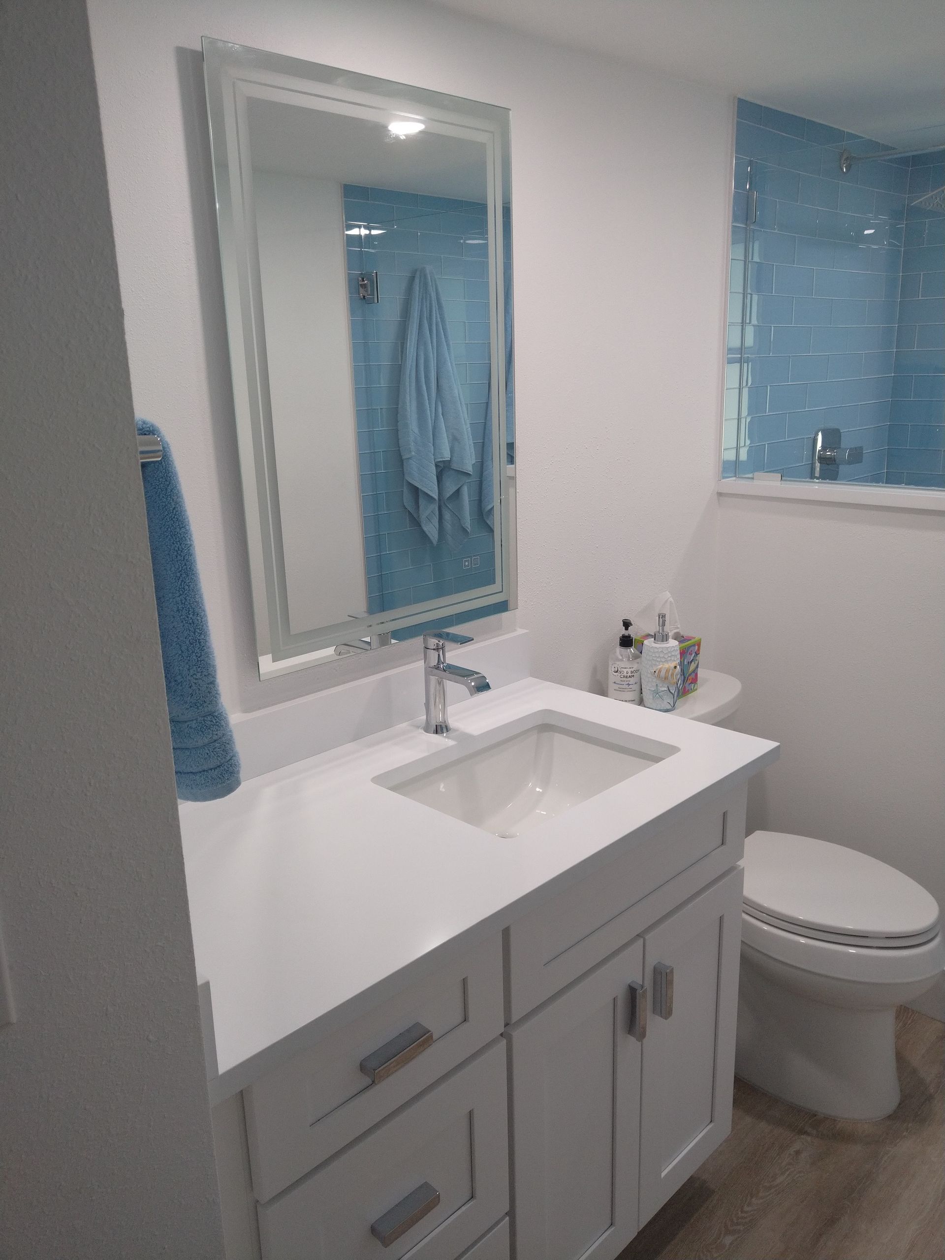 A white bathroom vanity with a square sink, faucet, and mirror, featuring blue towels and a blue-tiled shower in the back.