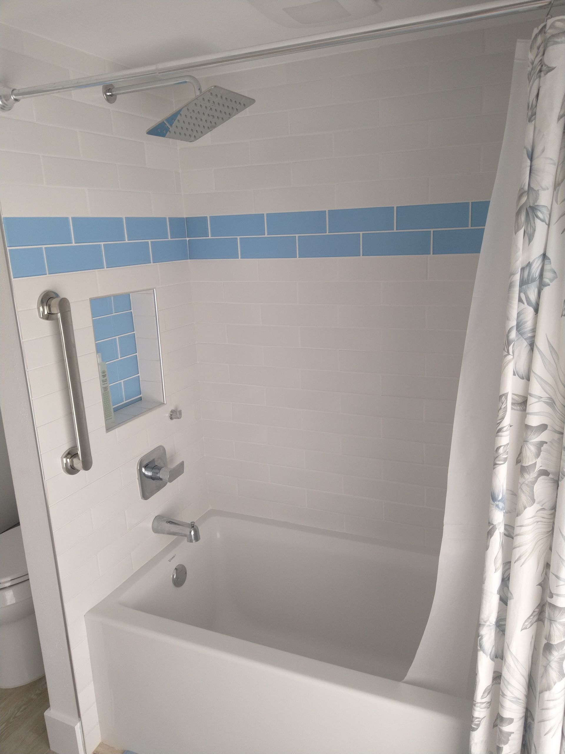 A white bathtub with a rainfall showerhead, a metal grab bar, and a blue tiled accent strip on the wall.