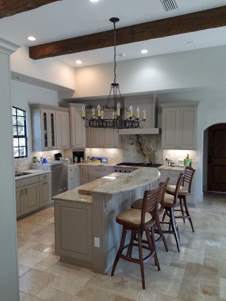 A kitchen with light cabinets, a curved island with wooden bar stools, stone-tile floors, and a wrought-iron chandelier.