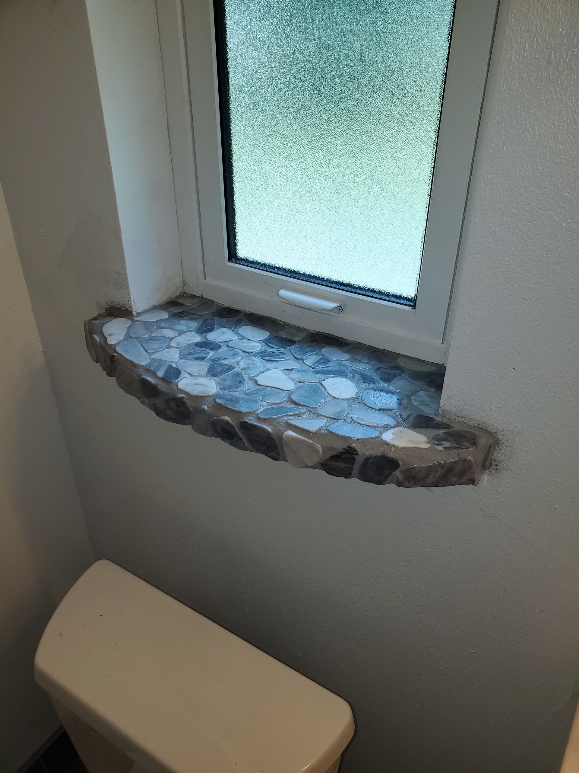 A pebble-tiled windowsill with a grey stone edge sits below a frosted window in a bathroom above a toilet tank.