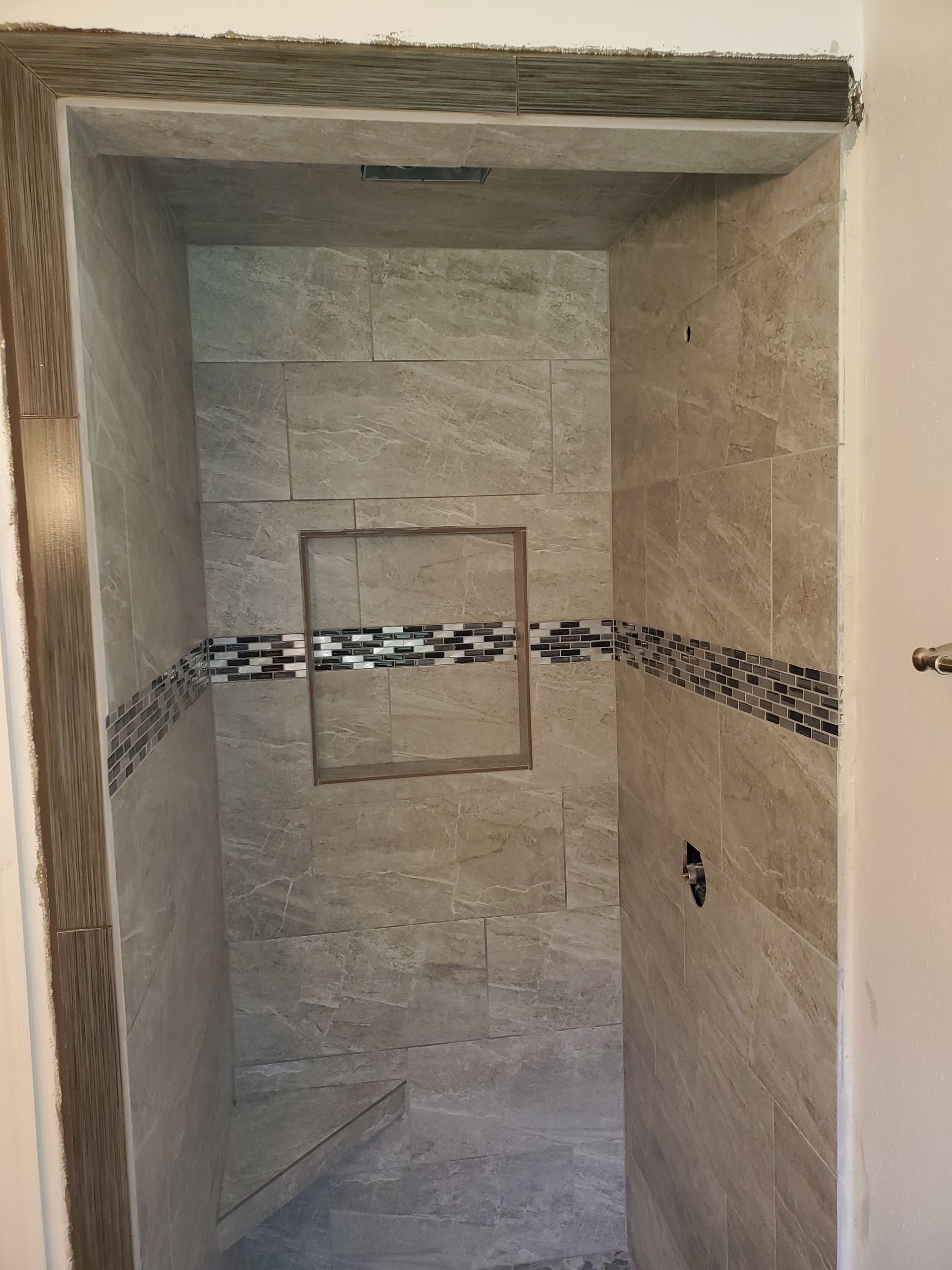A tiled walk-in shower under construction with a central wall niche and a horizontal decorative mosaic tile accent.