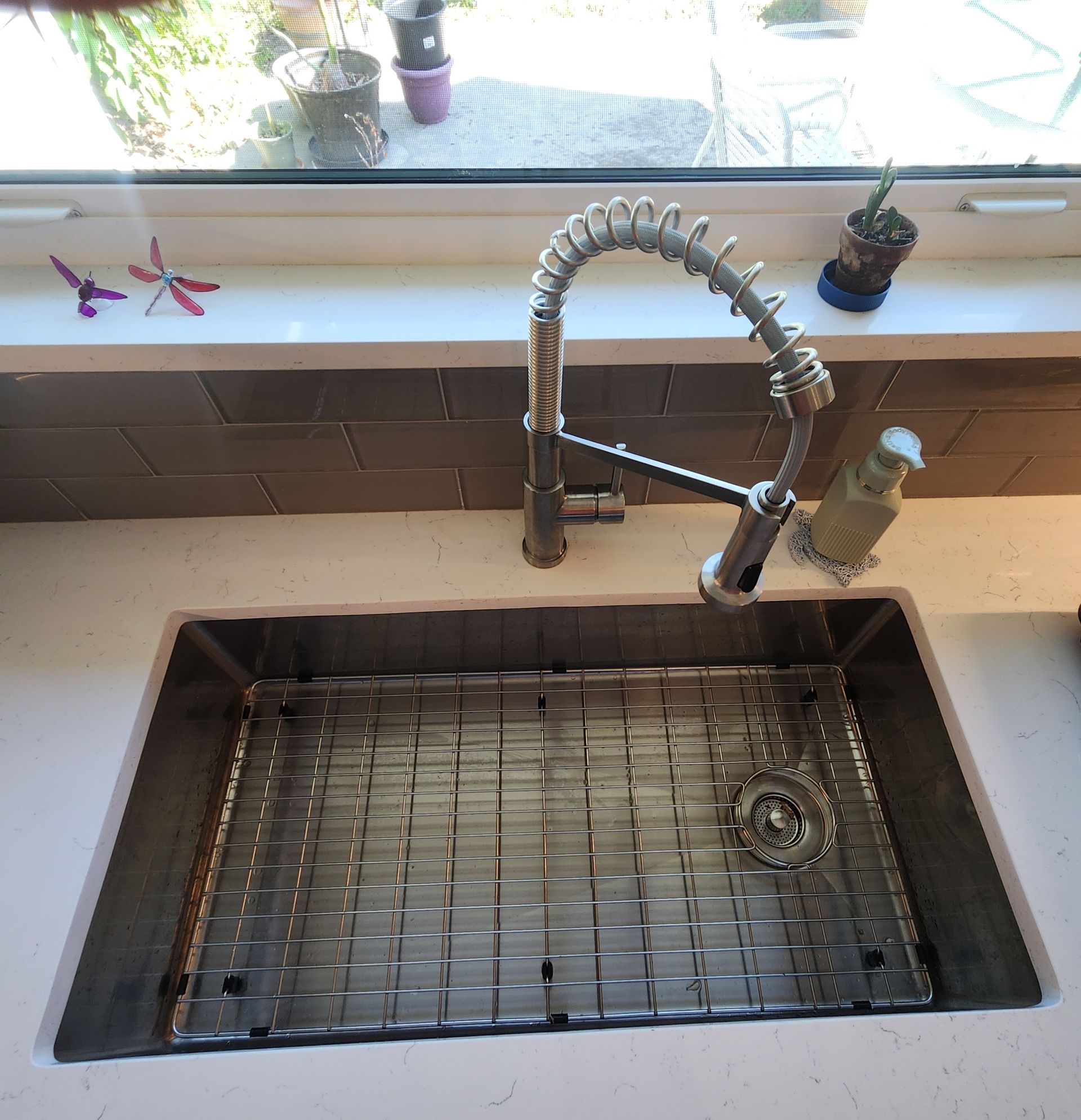 A stainless steel kitchen sink with a spring-spout faucet, a metal rack, and a soap dispenser against a tiled backsplash.