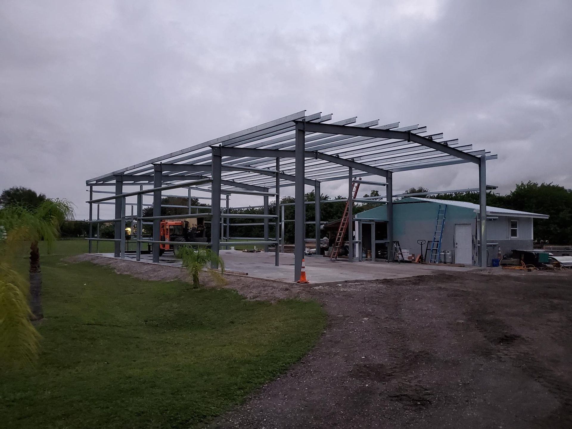A steel frame structure for a building under construction on a grassy lot next to a small house under cloudy skies.