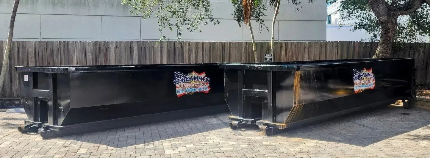 Two large black dumpsters sit side by side in front of a wooden fence, with a tree in the background.