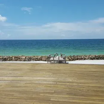 Wooden deck overlooking a beach with clear blue water and sky; small wooden seating area.