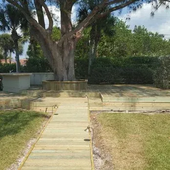 Wooden pathway leading to a large tree with tiered wooden platforms, surrounded by grass and foliage.