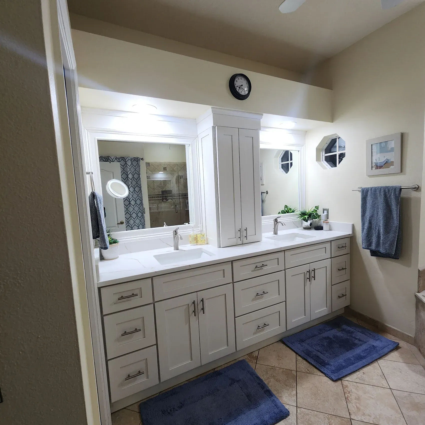 A white bathroom vanity with two sinks, a tall cabinet, and two blue rugs on a tile floor.