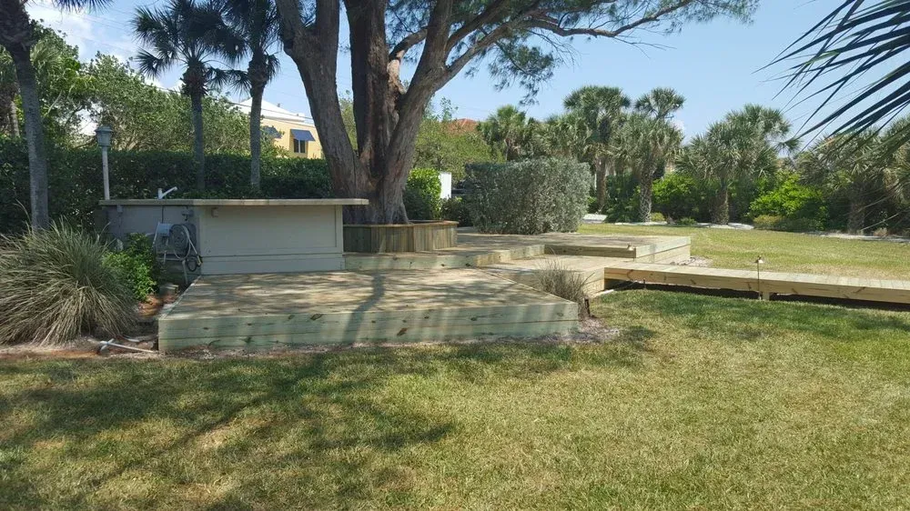 Outdoor space with tiered wooden platforms, grass, trees, and a light-colored structure.