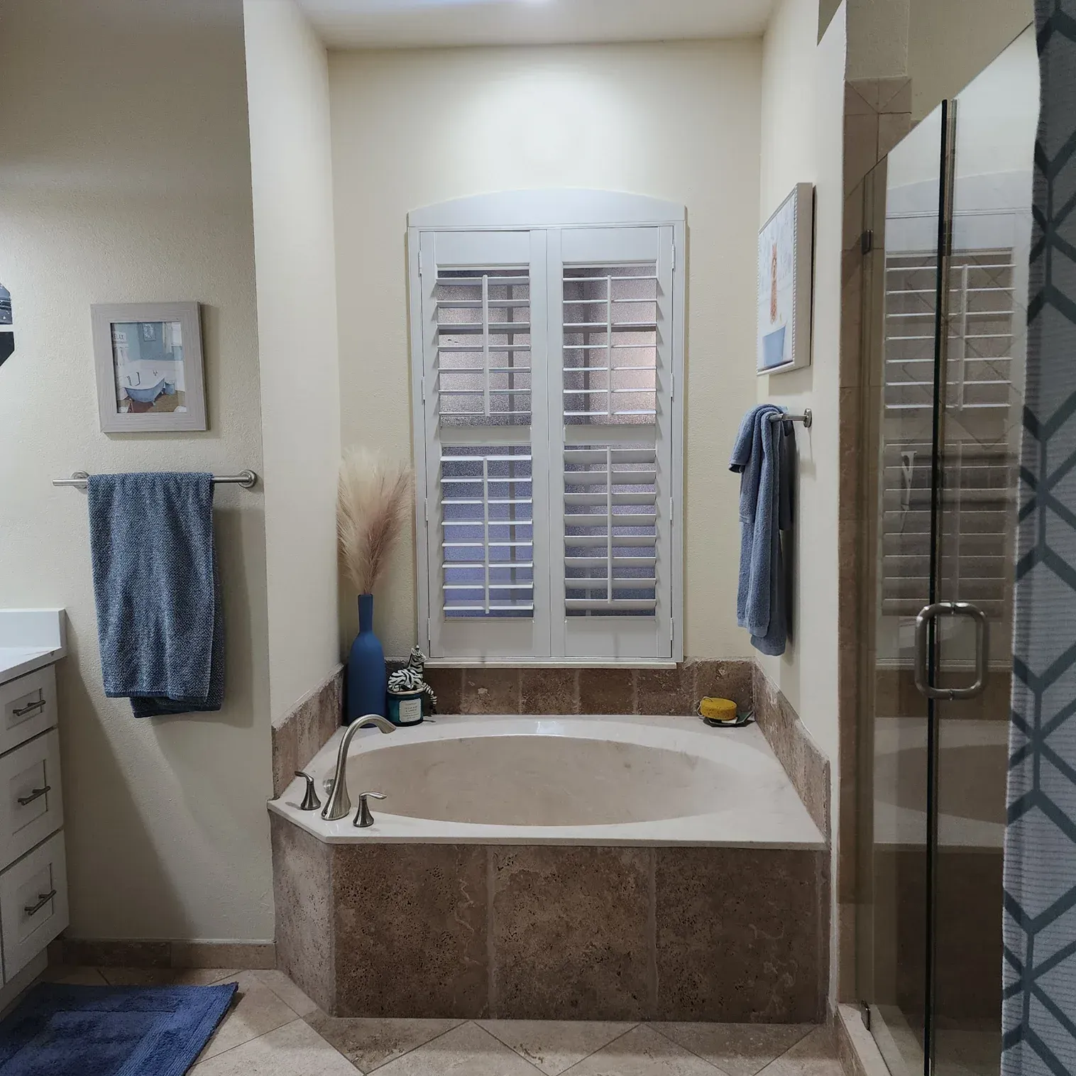 Bathroom with a corner bathtub, shower, and window with shutters. Beige walls and brown tile. Blue towels and rug.