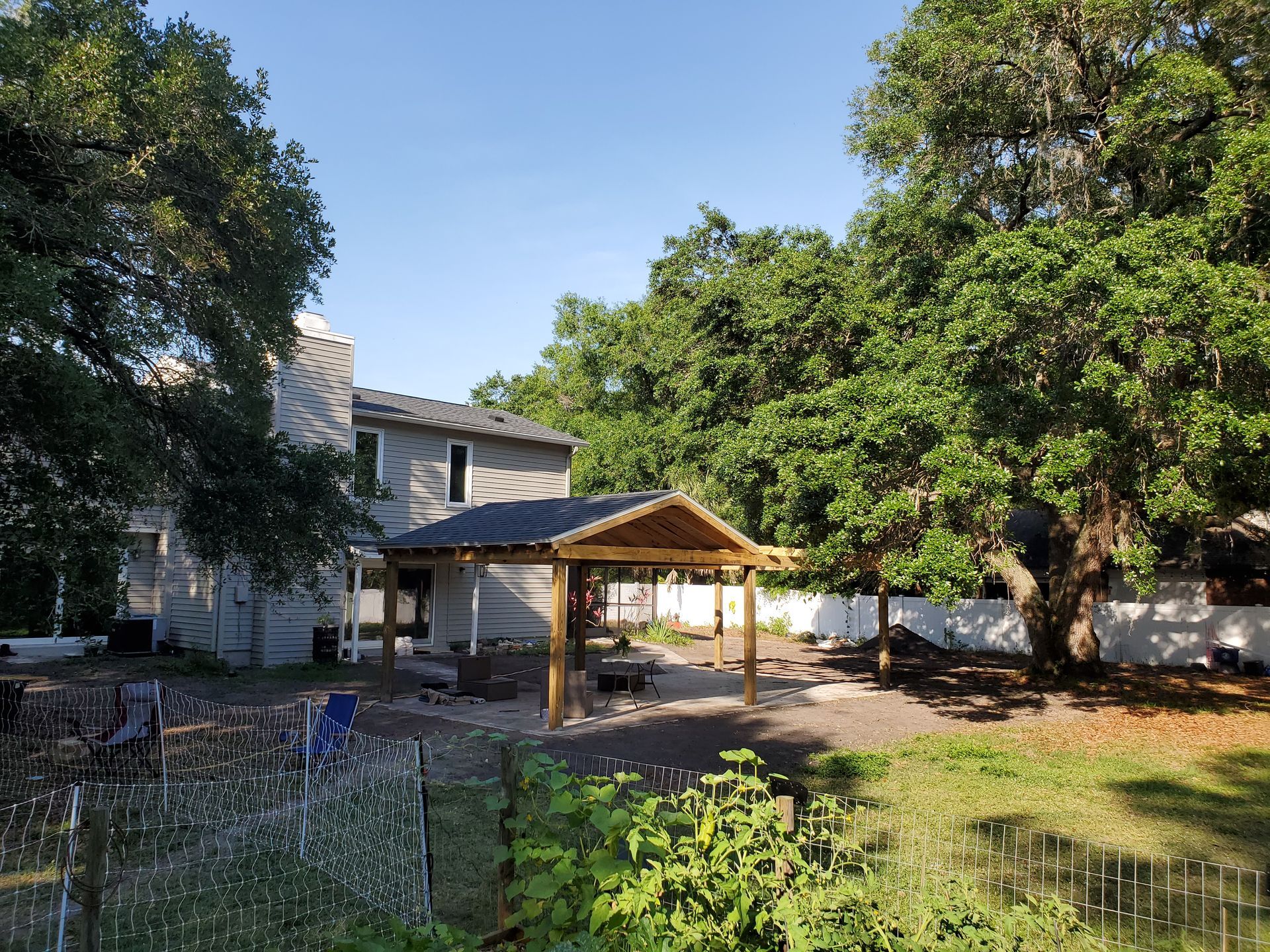 A backyard features a newly built wooden pergola on a stone patio next to a house, surrounded by large, leafy trees.