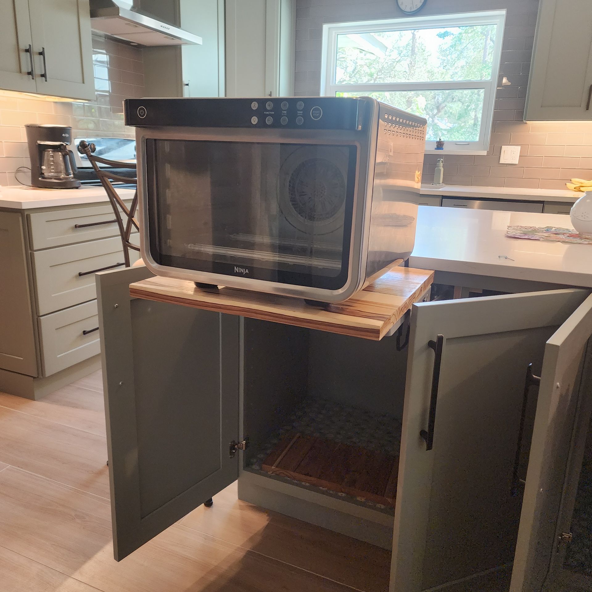 A countertop oven sits on a custom pull-out wooden board installed inside a lower kitchen cabinet with grey doors.