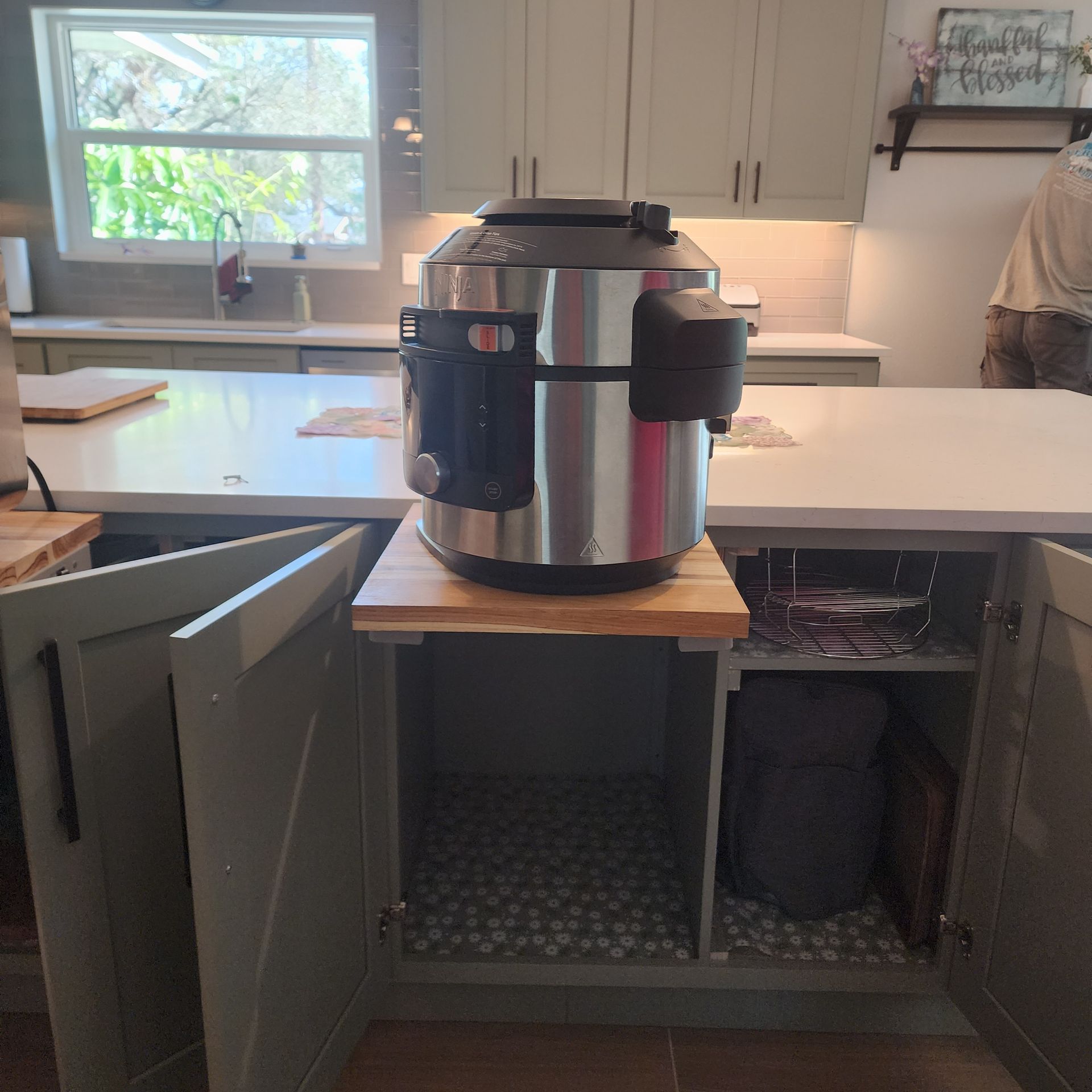 A stainless-steel multi-cooker sits on a wooden pull-out tray installed in an open kitchen island cabinet.
