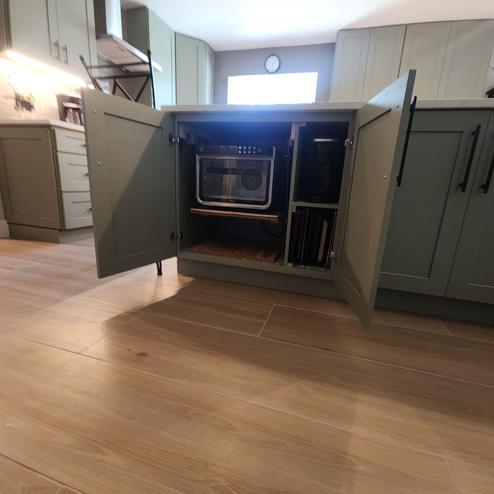 Sage green kitchen island cabinet open to reveal a microwave on a shelf, with vertical storage slots for pans nearby.