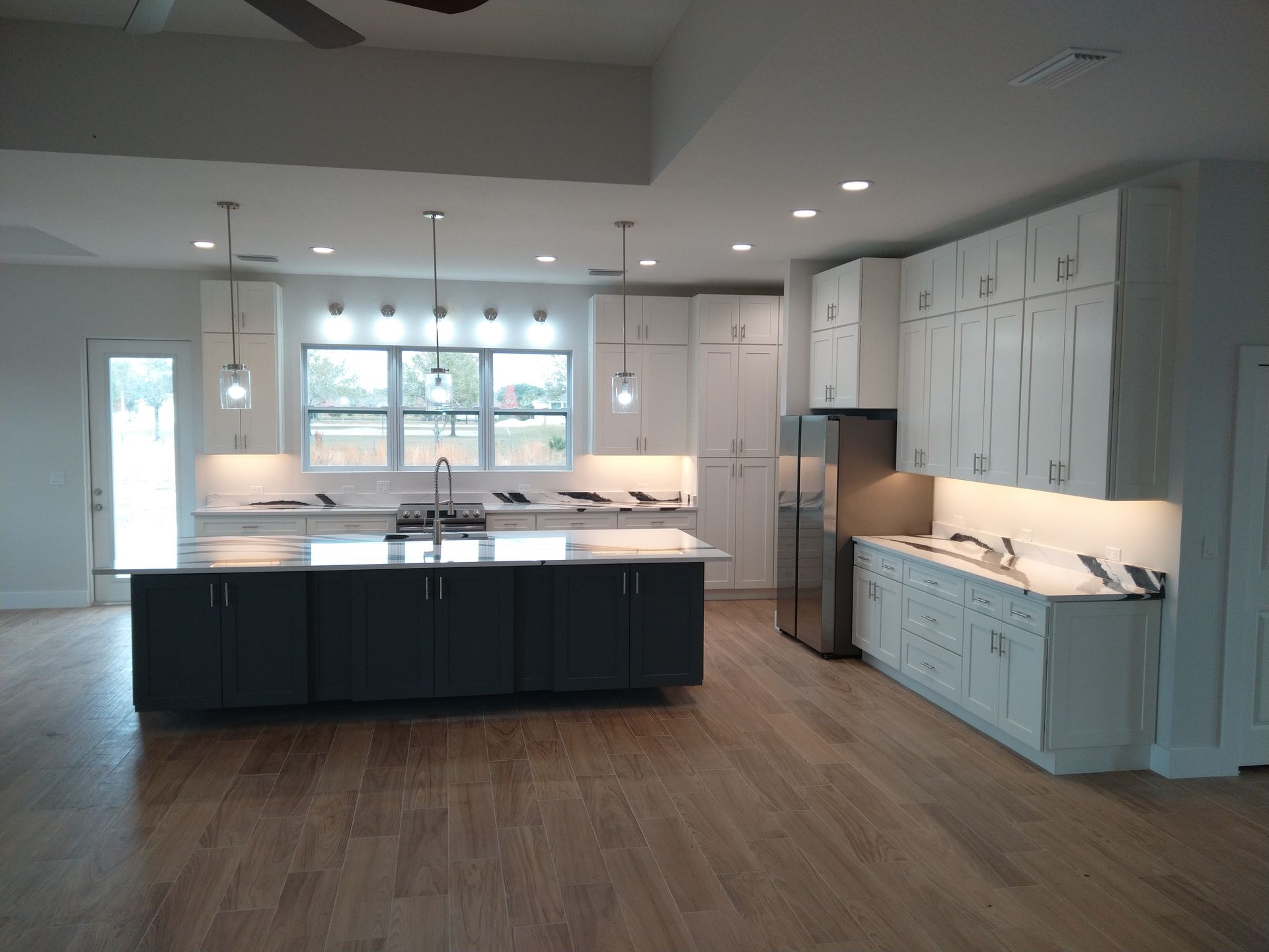A modern kitchen featuring a dark blue island, white cabinets, stainless steel appliances, and wood-look flooring.