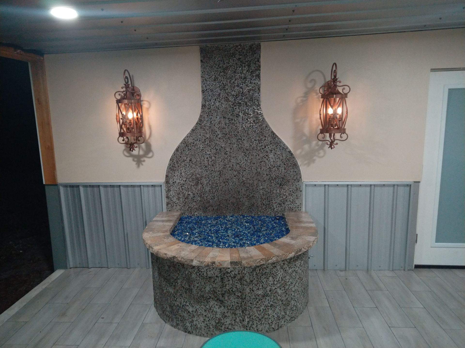 A semi-circular, stone-tiled fire pit with a tall, decorative stone backsplash, flanked by two ornate wall-mounted lamps.