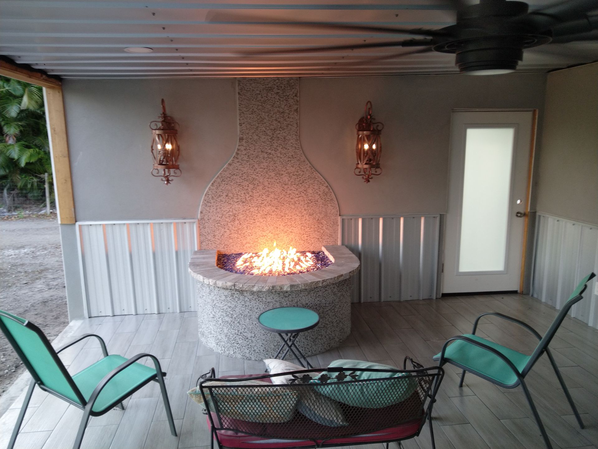 A covered patio featuring a central stone fire pit, two green chairs, and a small bench set against a neutral-toned wall.