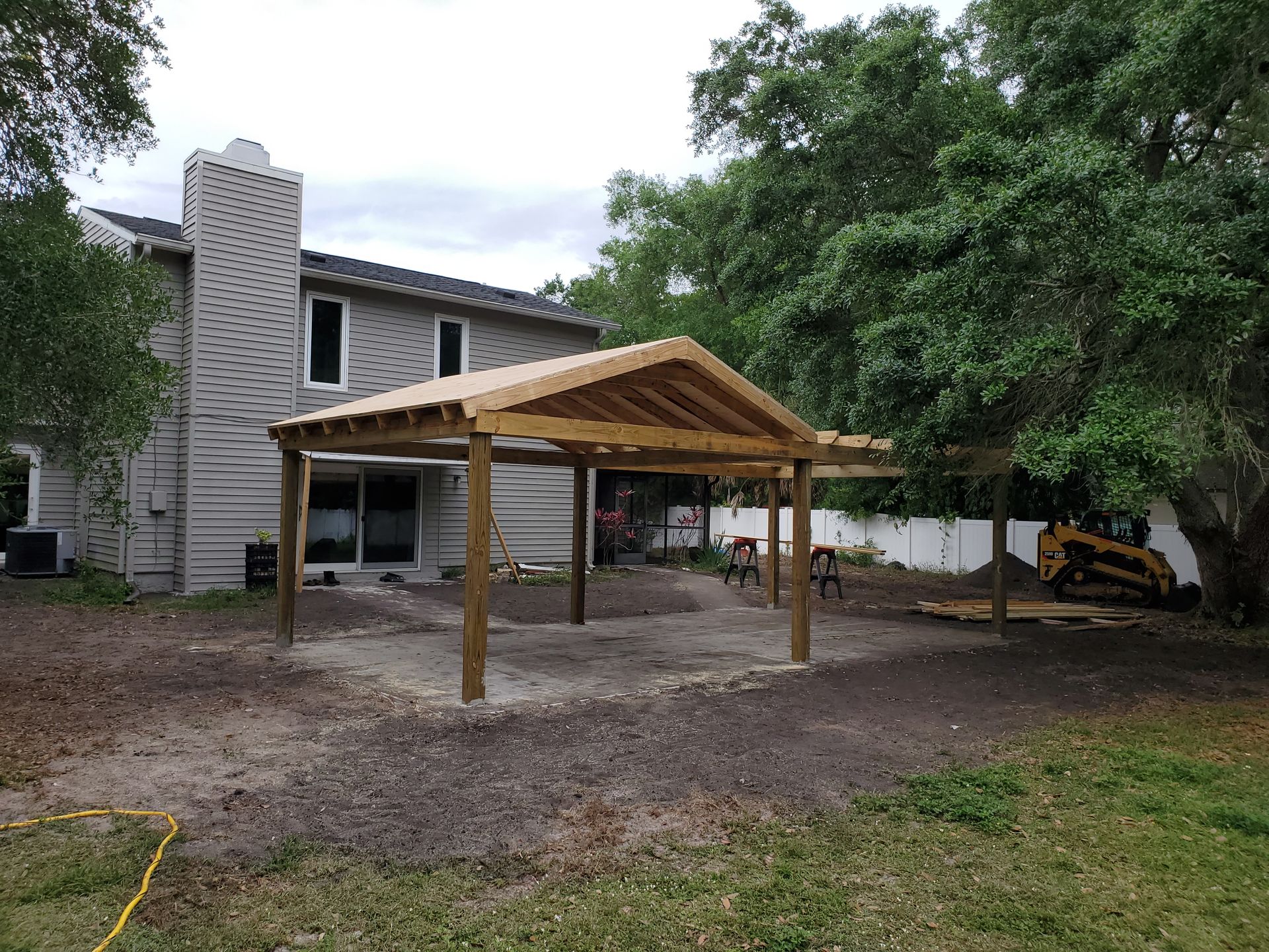 A newly constructed wooden patio cover stands over a concrete slab in the backyard of a two-story gray house.