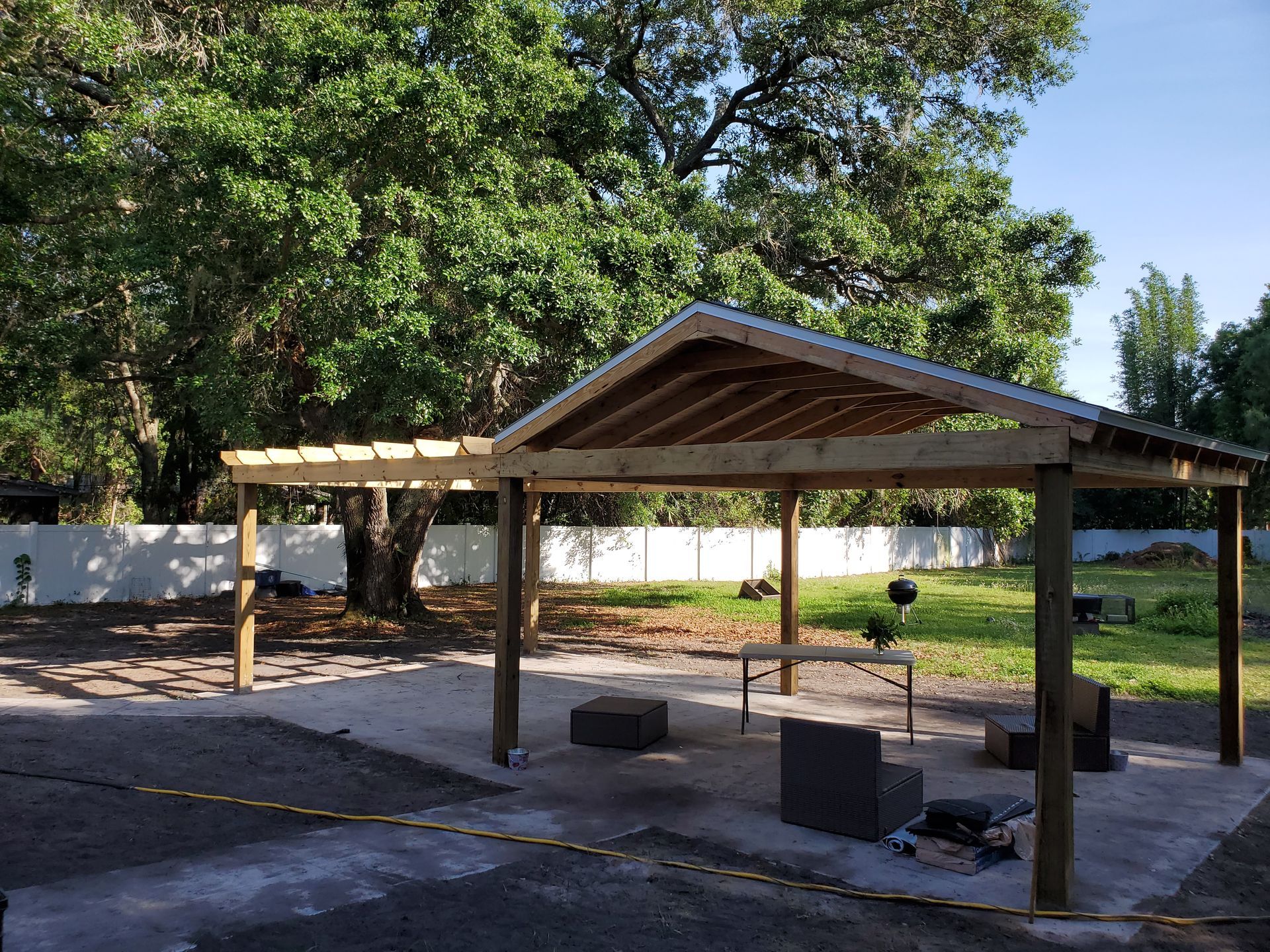 A partially covered wooden patio structure with a metal roof and open pergola section stands on a concrete slab in a yard.