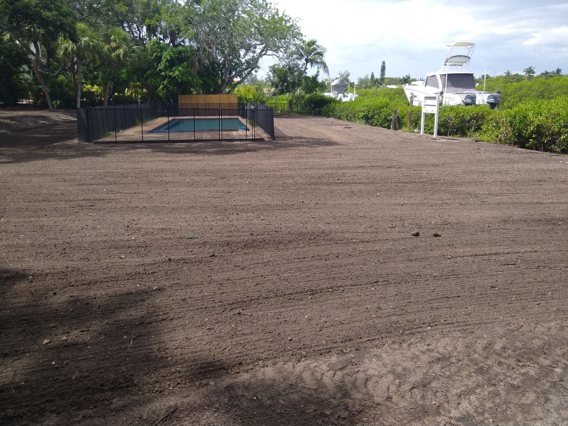 A dirt yard with a fenced-in swimming pool in the center, surrounded by trees and a boat parked in the background.