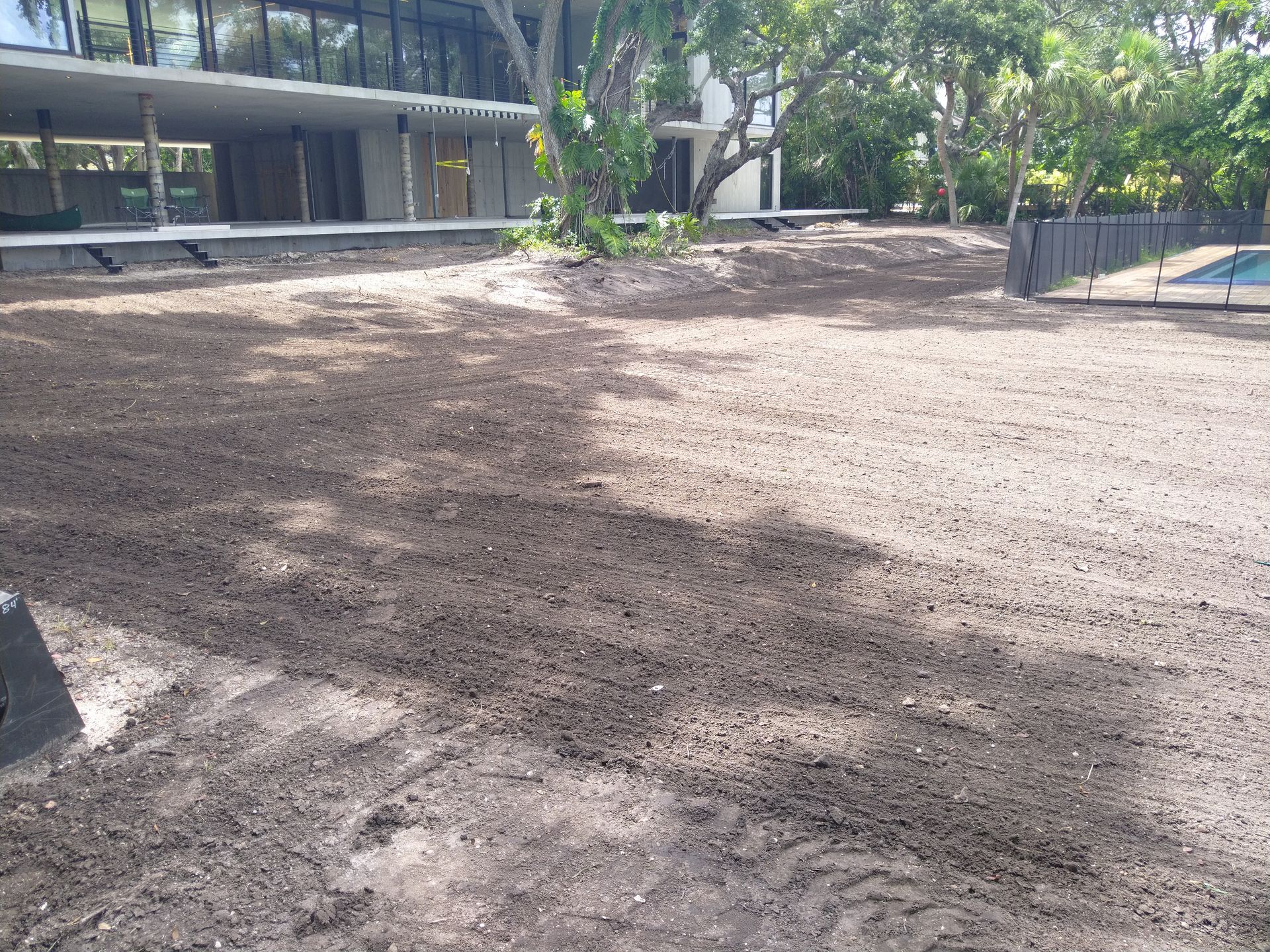 A large, freshly leveled patch of dark, tilled soil in a backyard next to a modern building and trees.