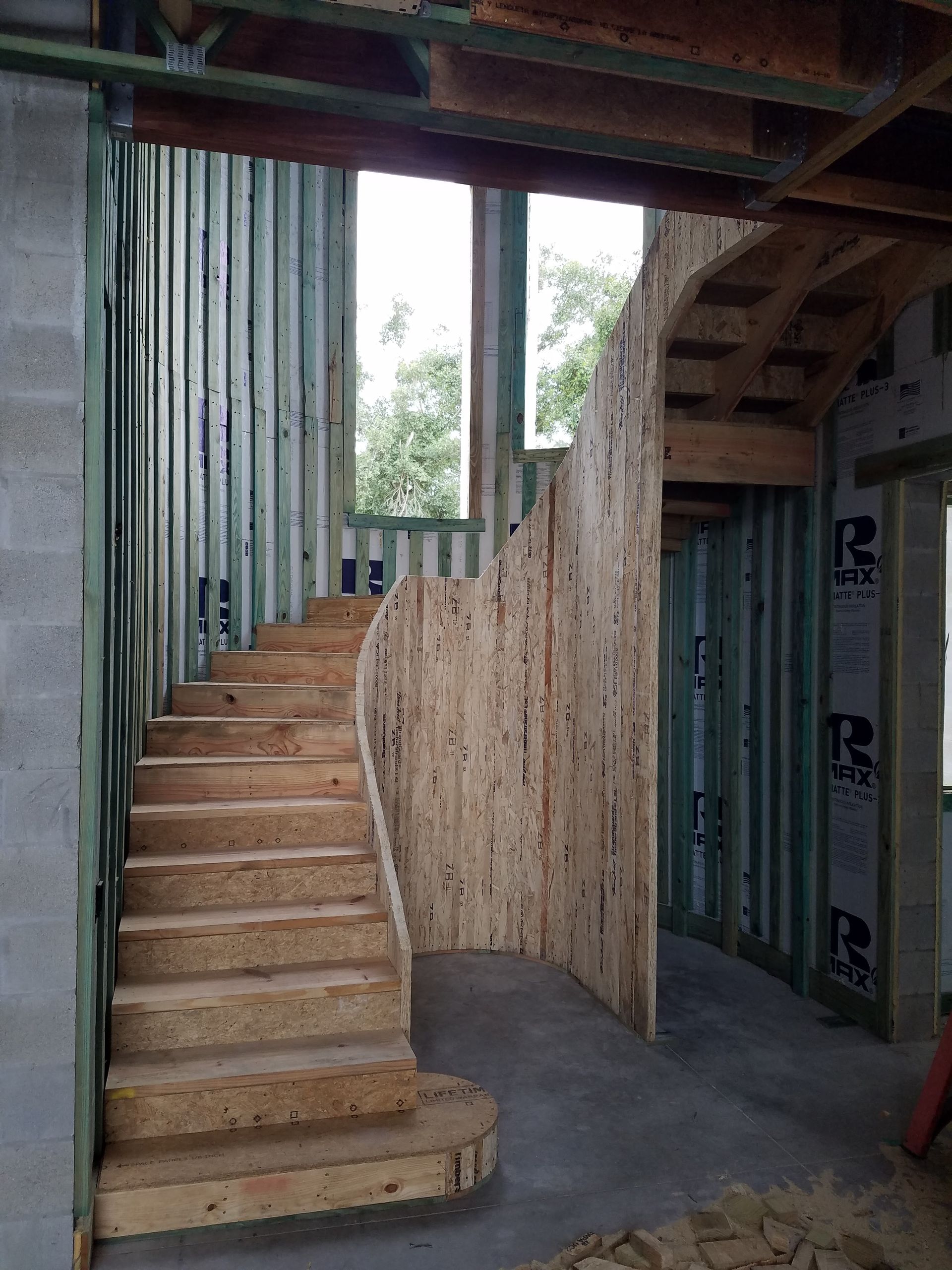 Unfinished wooden curved staircase in a building under construction with exposed metal wall studs and concrete floor.