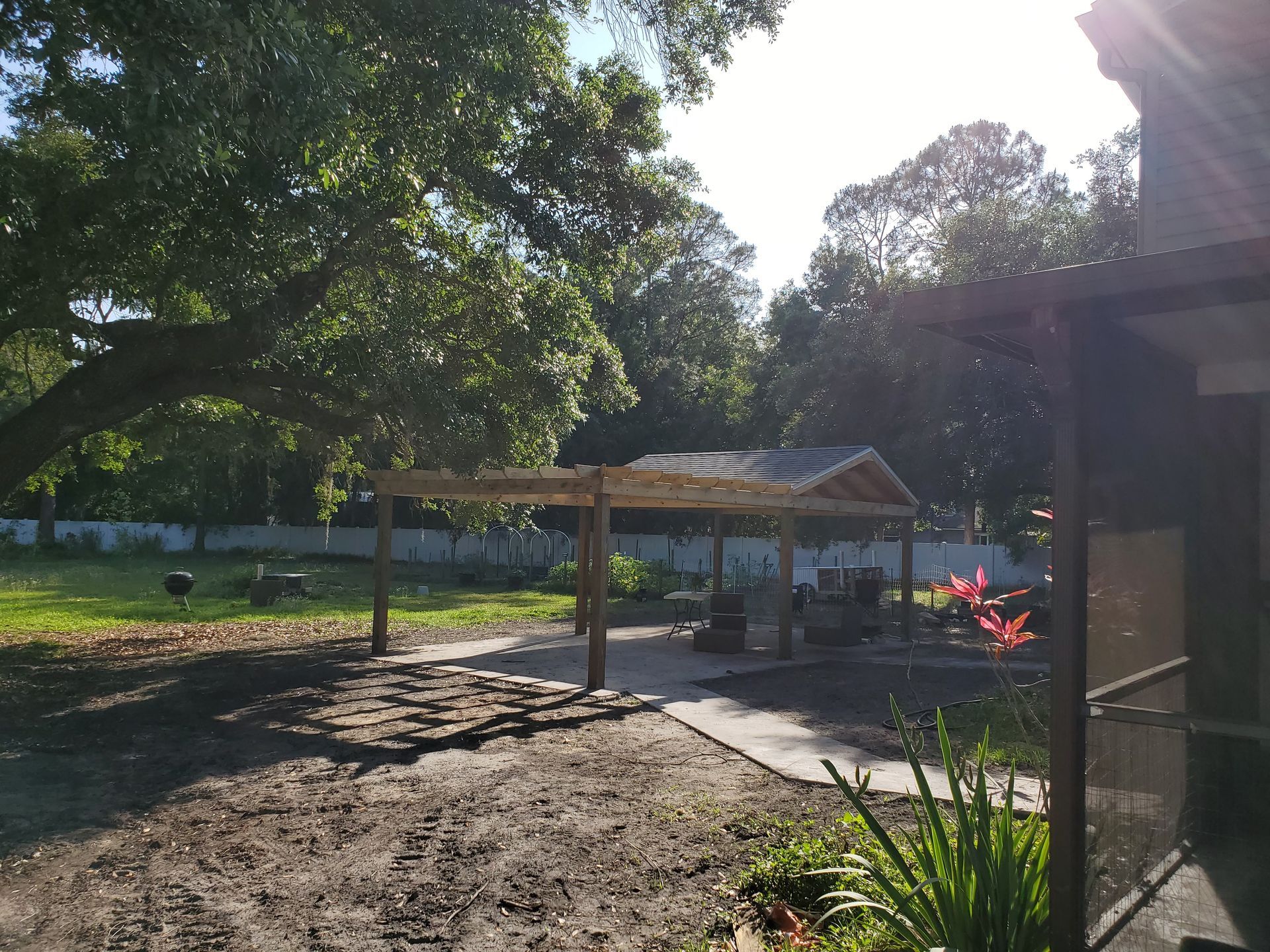 A wooden pergola stands on a paved patio in a grassy backyard near a house, surrounded by large, leafy trees.