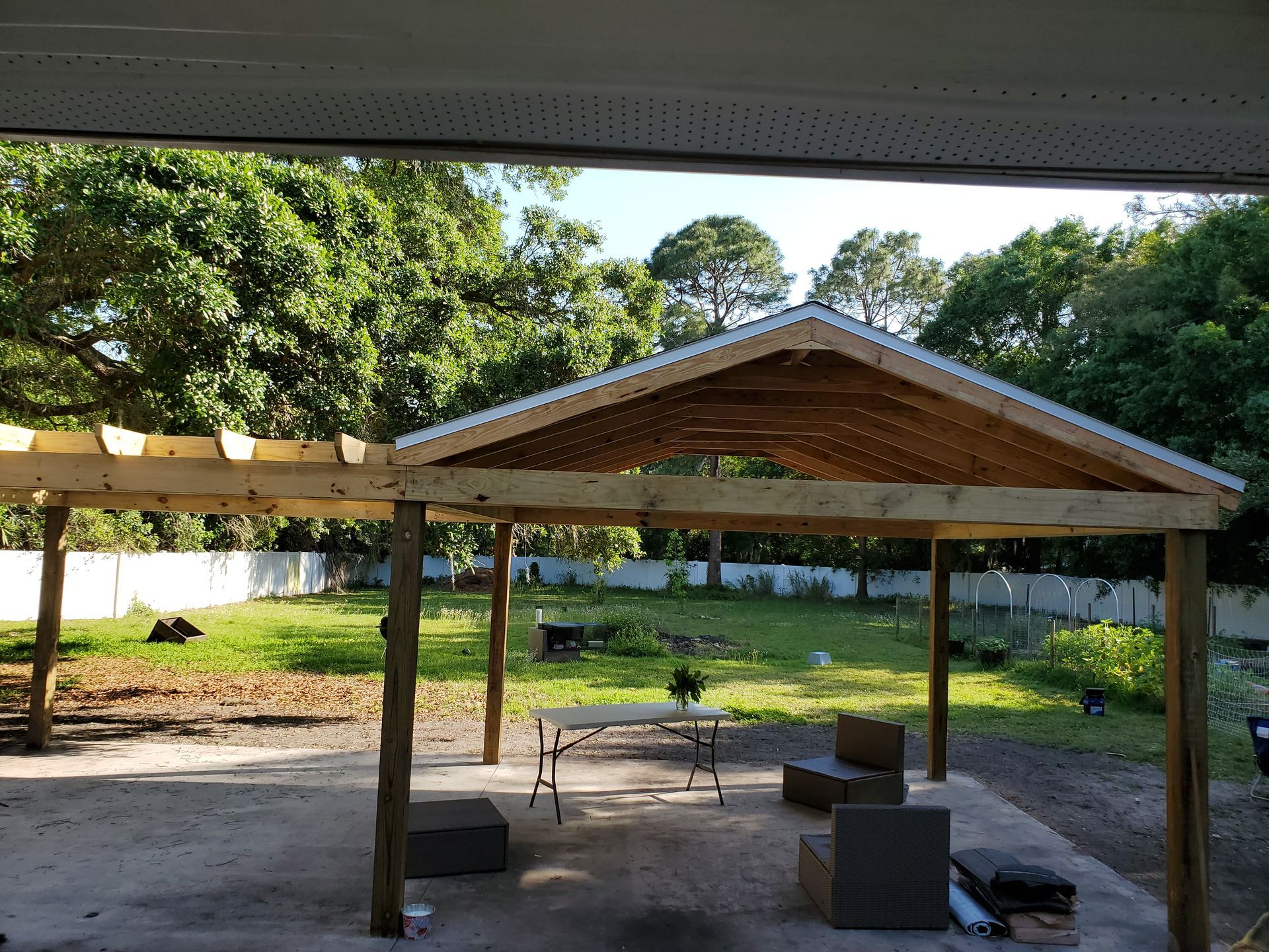 A partially constructed wooden pavilion structure with a metal roof over a concrete patio in a grassy backyard.