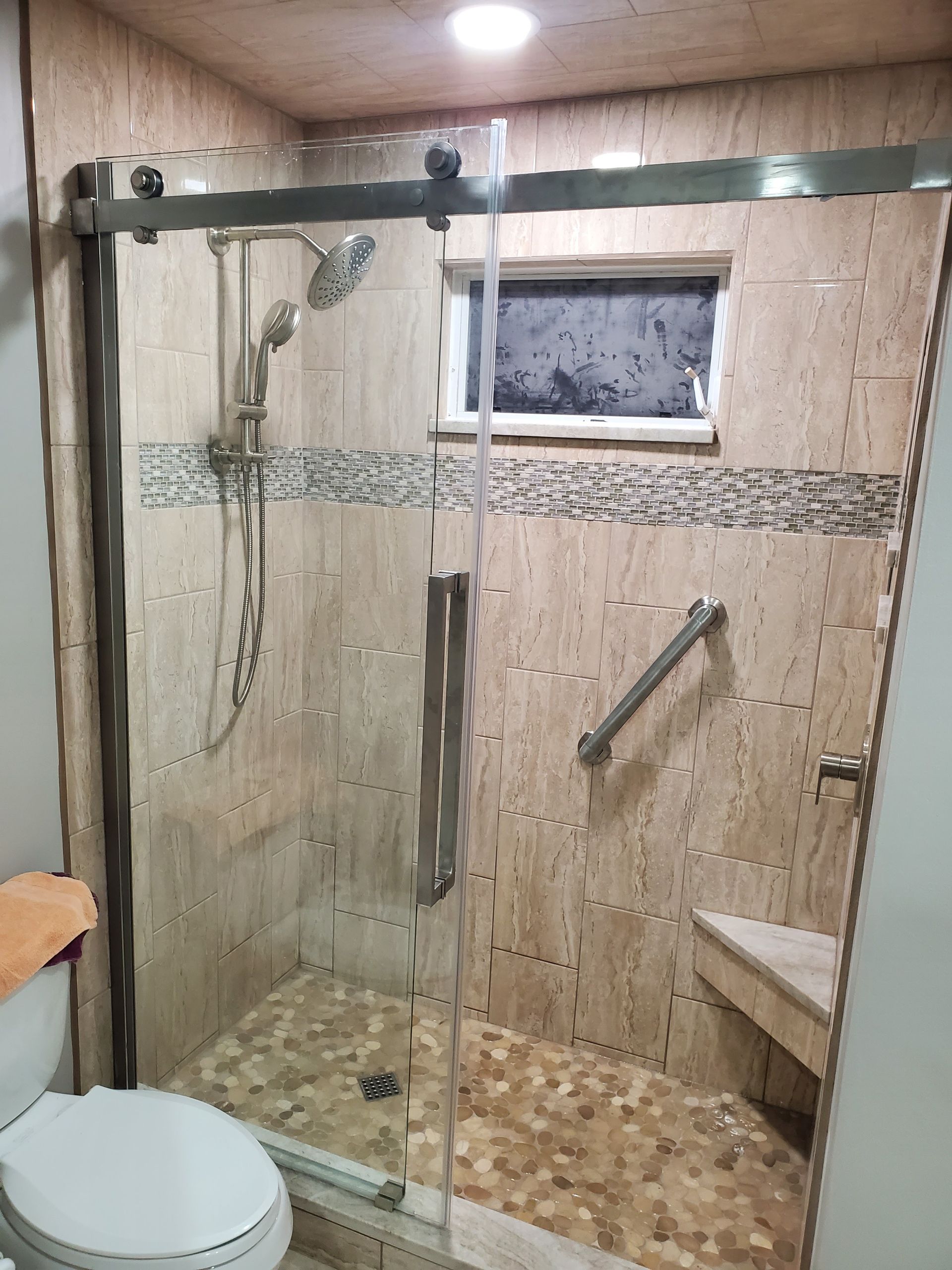 A glass-enclosed shower with beige tiled walls, a built-in seat, a grab bar, and a handheld shower head next to a toilet.