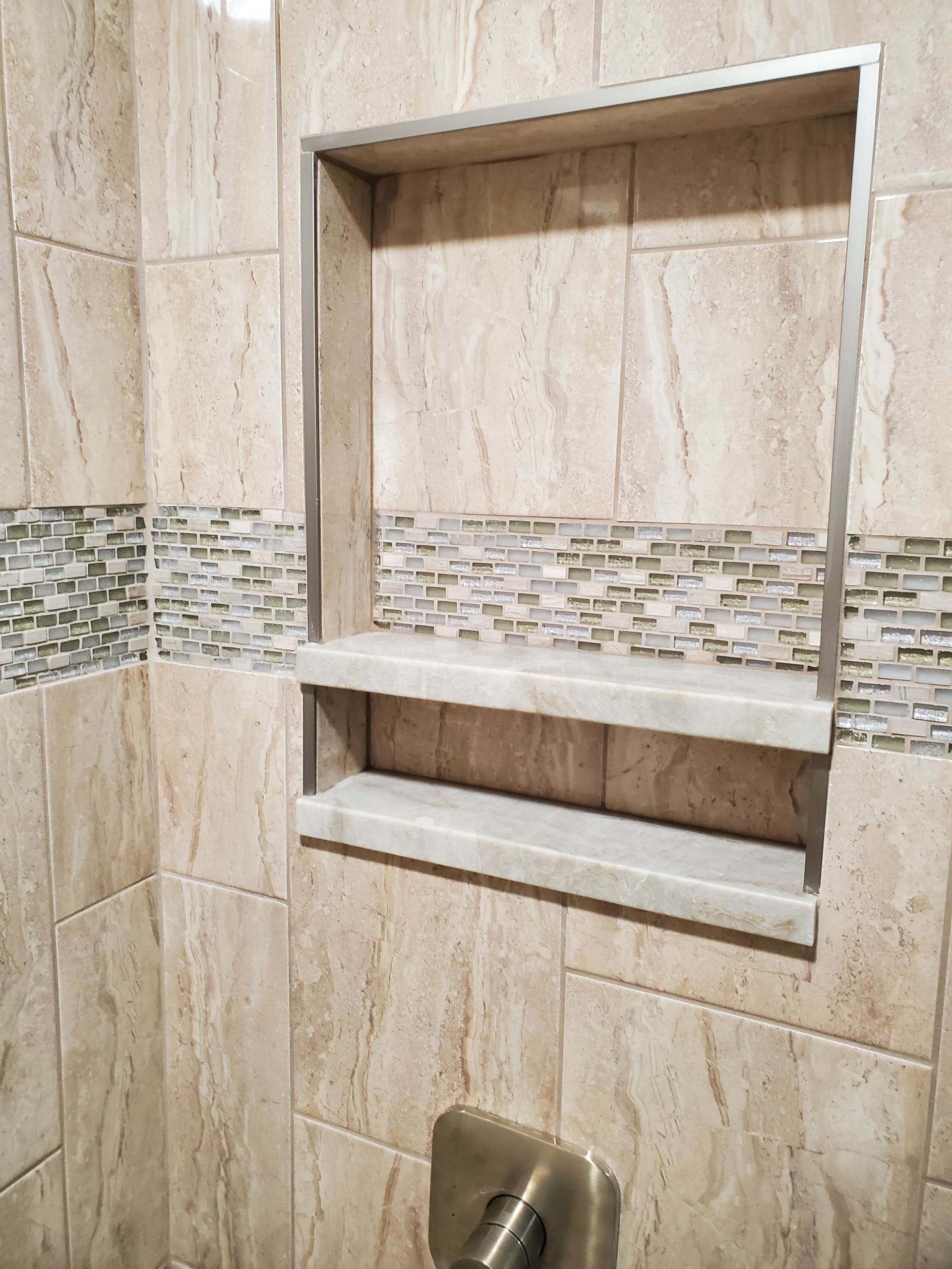 A beige tiled shower wall with a recessed storage niche containing two stone shelves and a metallic trim accent border.