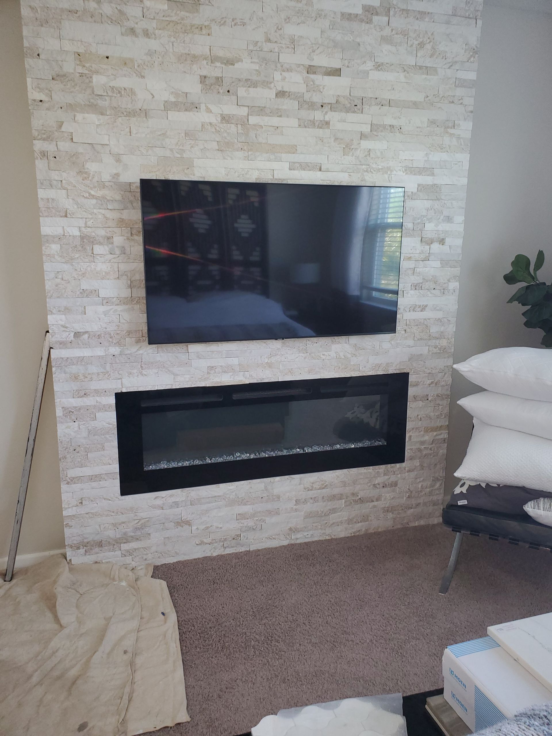A wall-mounted TV sits above a horizontal electric fireplace on a beige stone-tiled accent wall in a room.