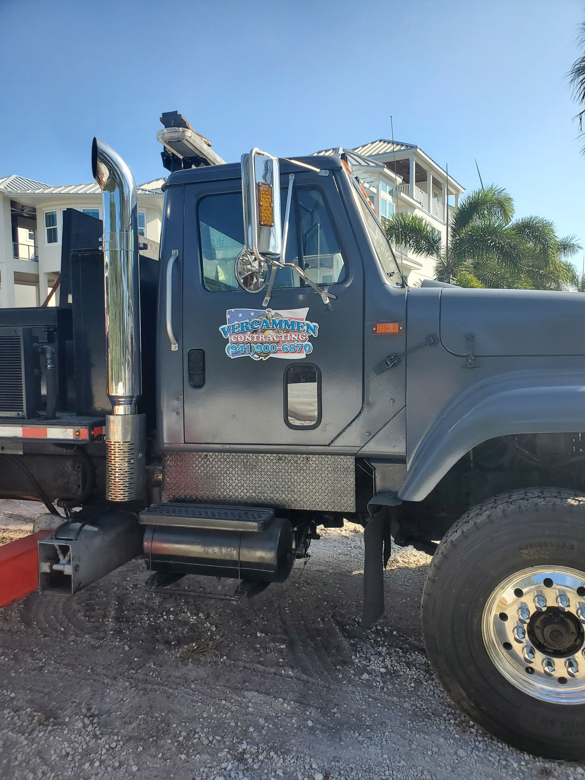 A dark gray heavy-duty truck parked on a gravel lot, featuring a large chrome exhaust stack and a custom door logo.