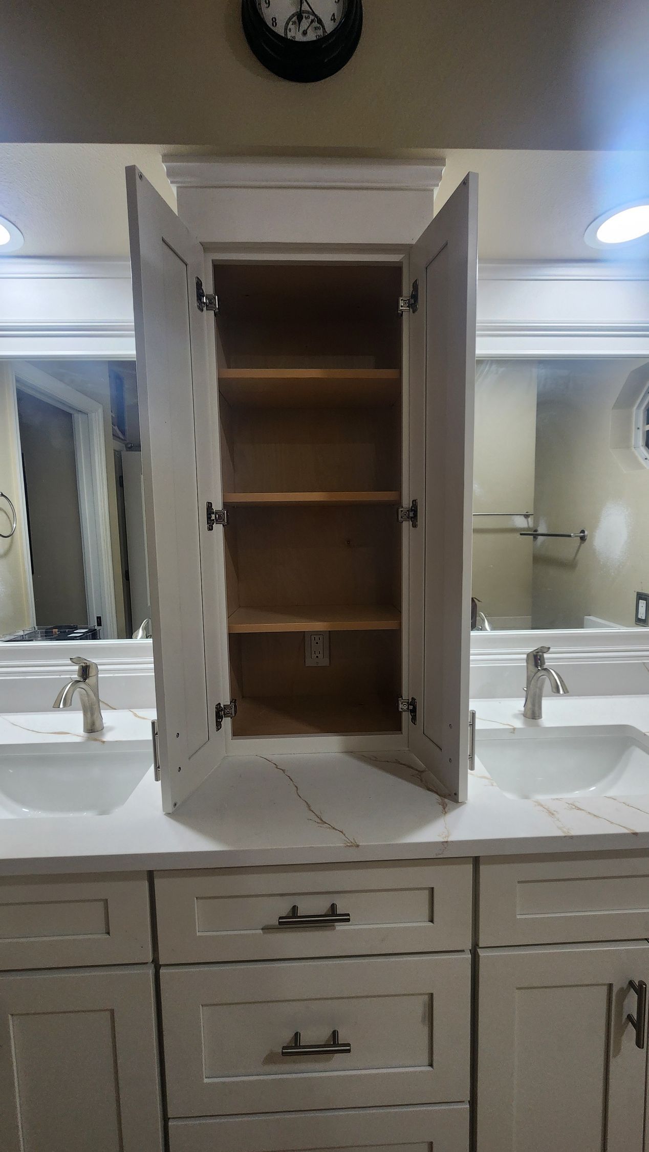 A white bathroom vanity with a central cabinet featuring open double doors and three wooden shelves, between two sinks.