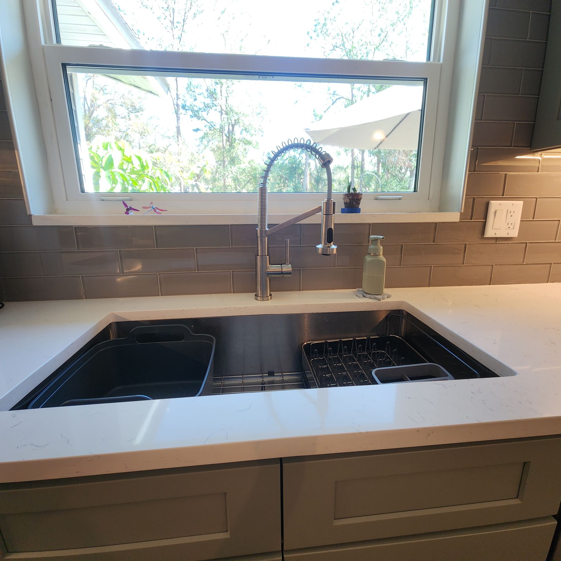 A stainless steel kitchen sink with a gooseneck faucet, set in a white countertop against a gray tiled backsplash.