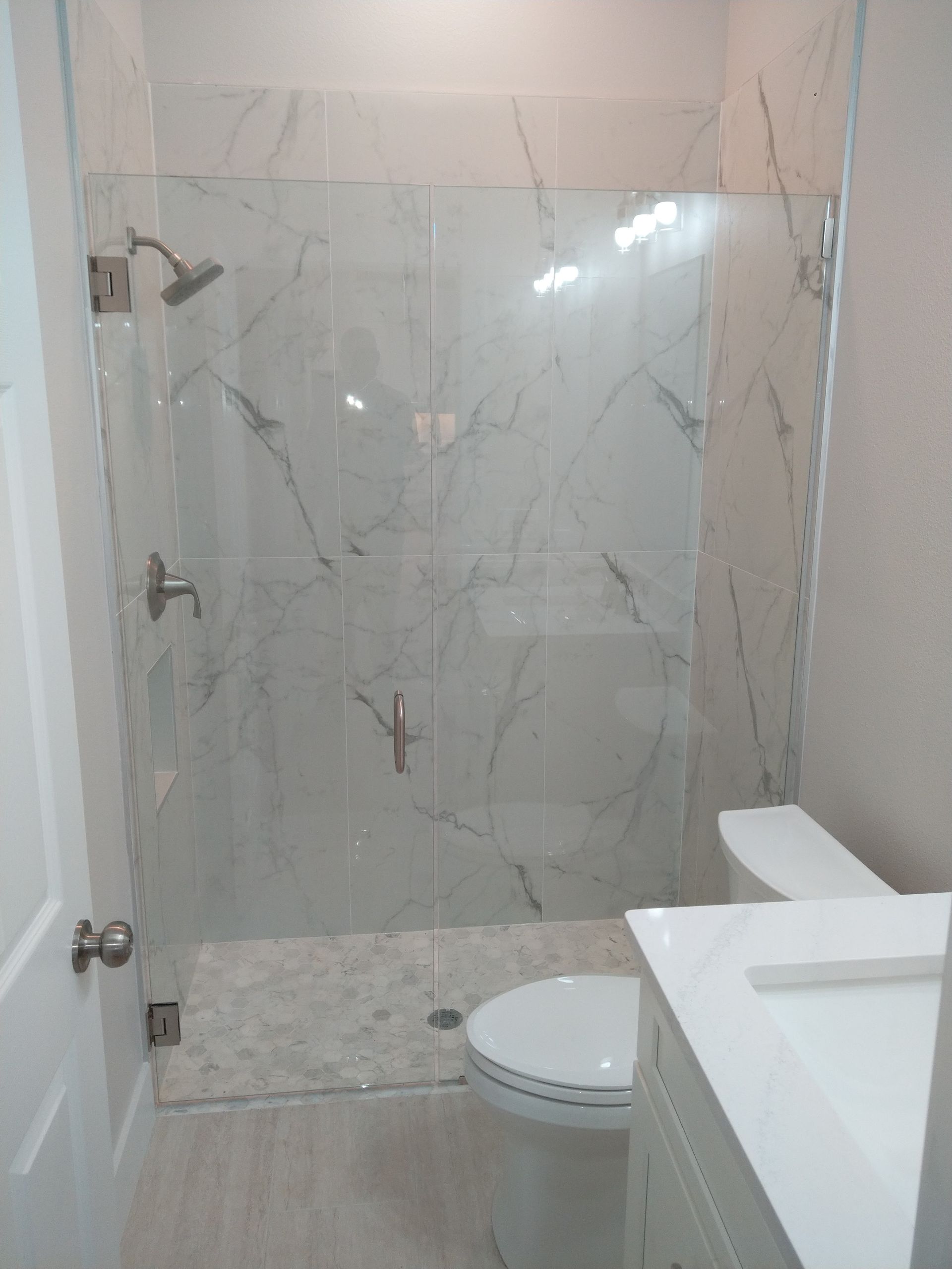 A modern bathroom featuring a white marble-tiled walk-in shower with a glass door, next to a white toilet and vanity.