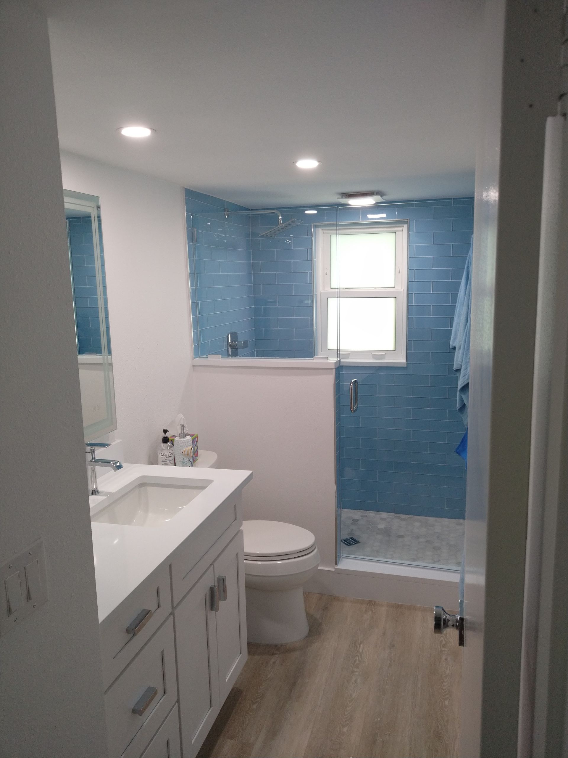 A bathroom with a white vanity, a light wood floor, a white toilet, and a walk-in shower with blue tiled walls.