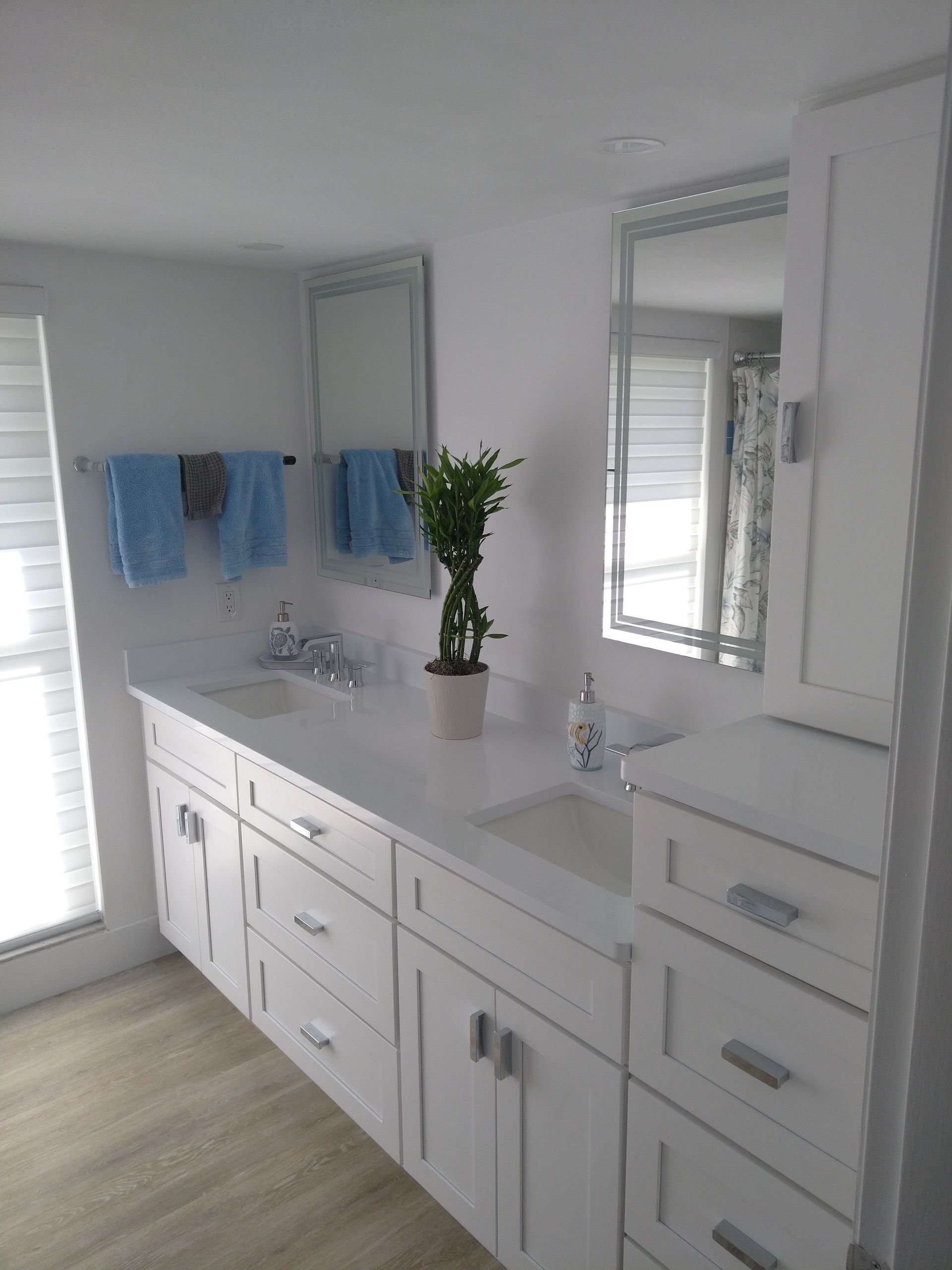 Modern bathroom vanity with white cabinets, double sinks, two mirrors, and a potted plant in a bright, white room.