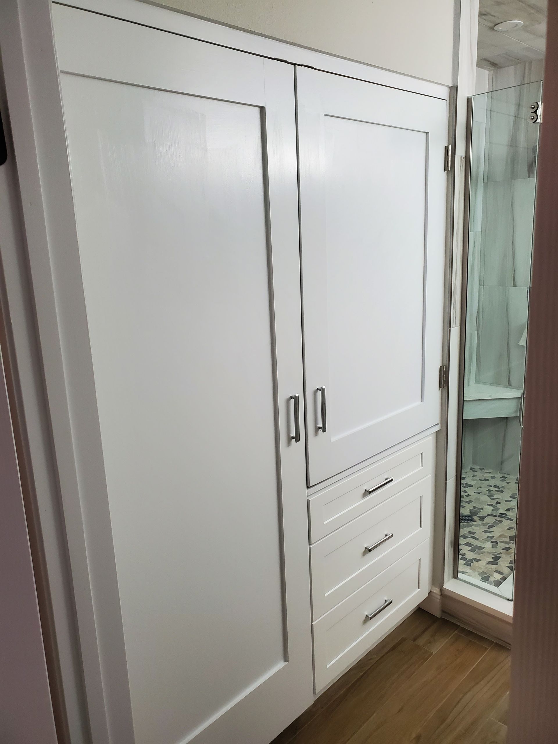 A floor-to-ceiling white cabinet with two doors and three drawers next to a bathroom with a glass shower door.