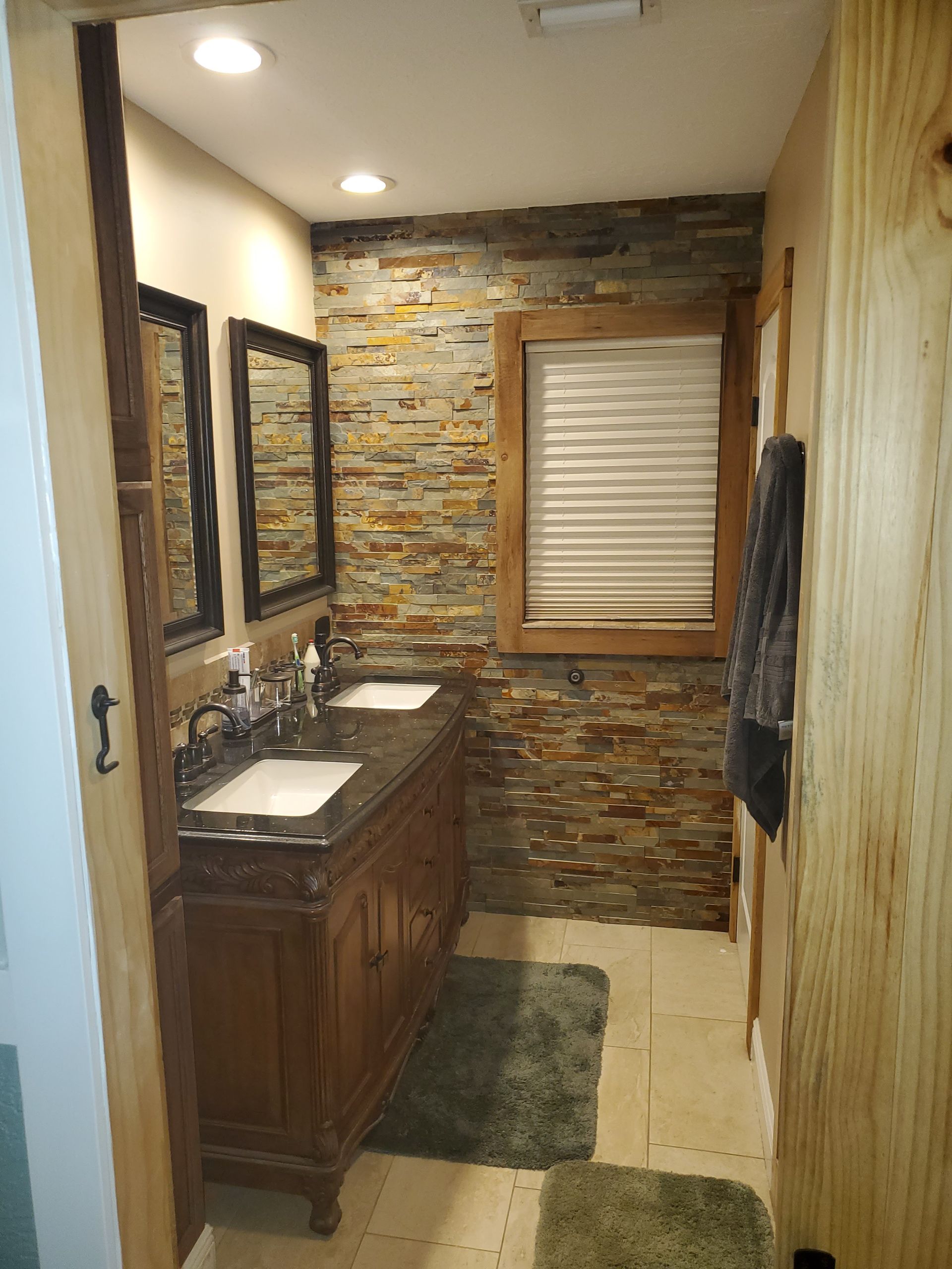 A rustic bathroom featuring a stone accent wall, wooden vanity with double sinks, two framed mirrors, and tiled flooring.