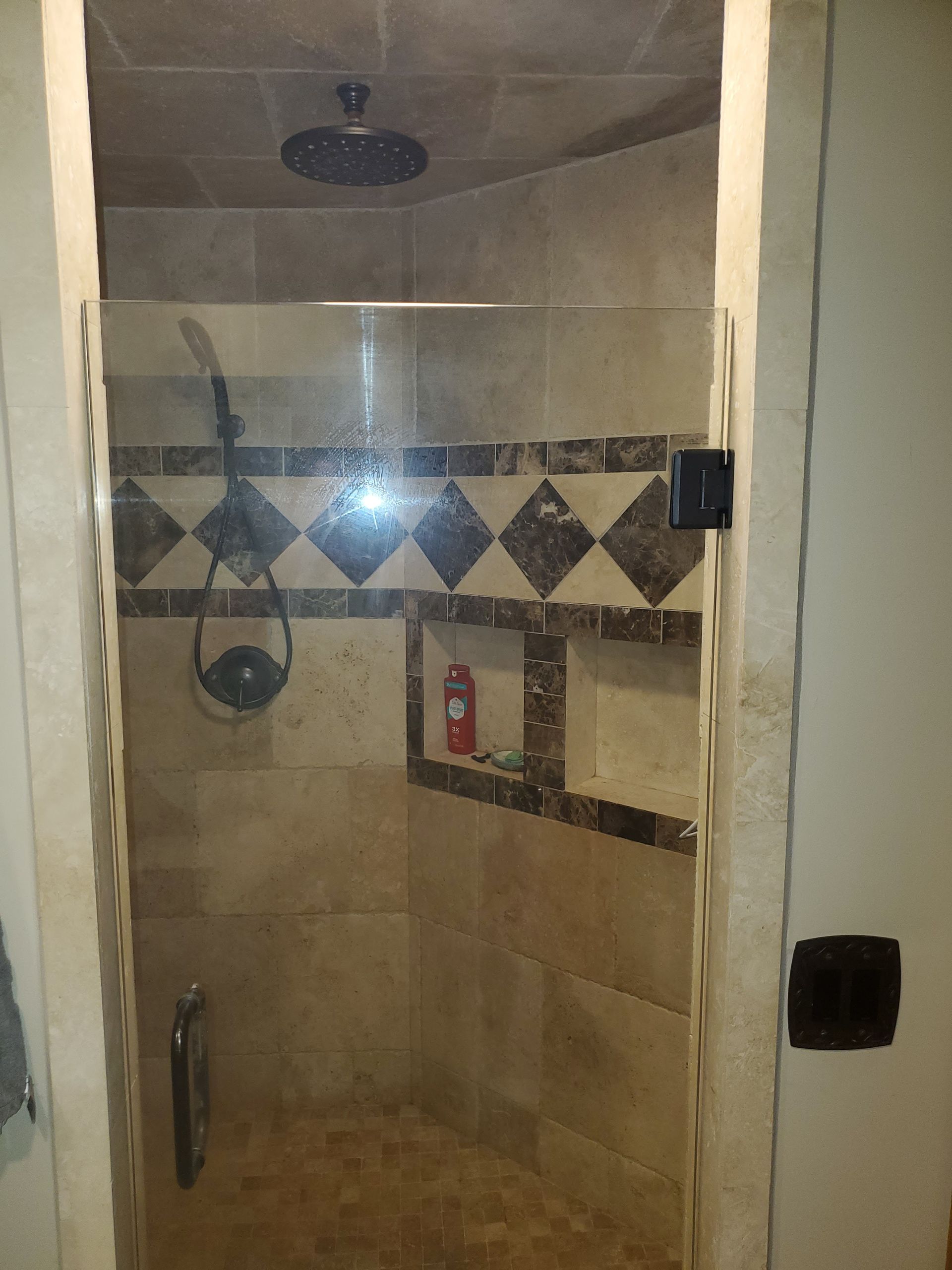 A walk-in shower with tan tiled walls, a decorative dark diamond-pattern border, a glass door, and a built-in shelf.