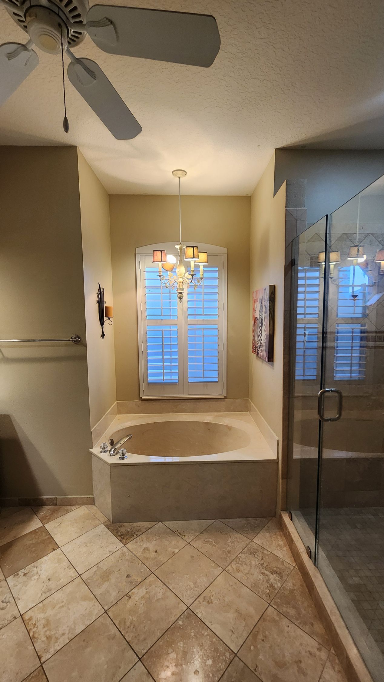A bathroom with a built-in tub under a window, a ceiling fan, and a glass-enclosed shower on the right.